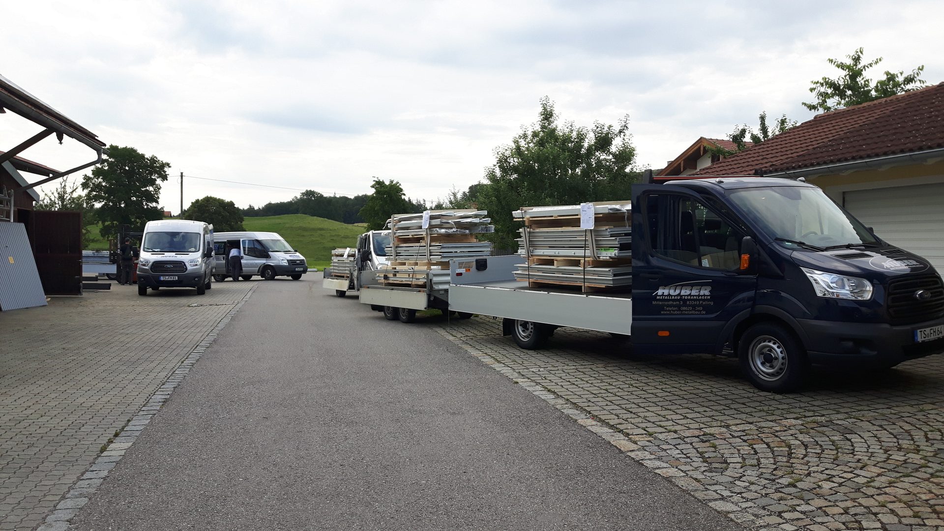 Vans and flatbed trailers loaded with materials, likely for a metal construction company, parked on a road.