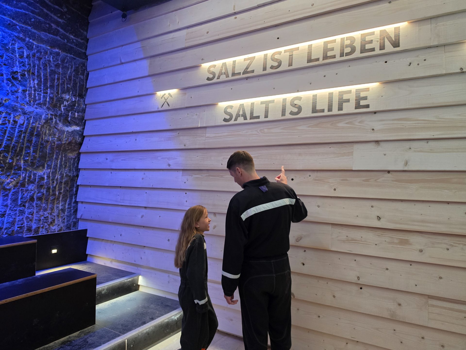 Man and child look at a wooden wall displaying "Salt is Life" in German and English, with blue lighting.