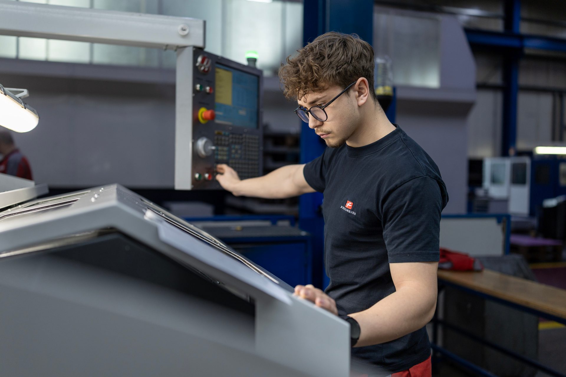 A young man in glasses operates a CNC machine control panel in a workshop, focusing intently on the task.