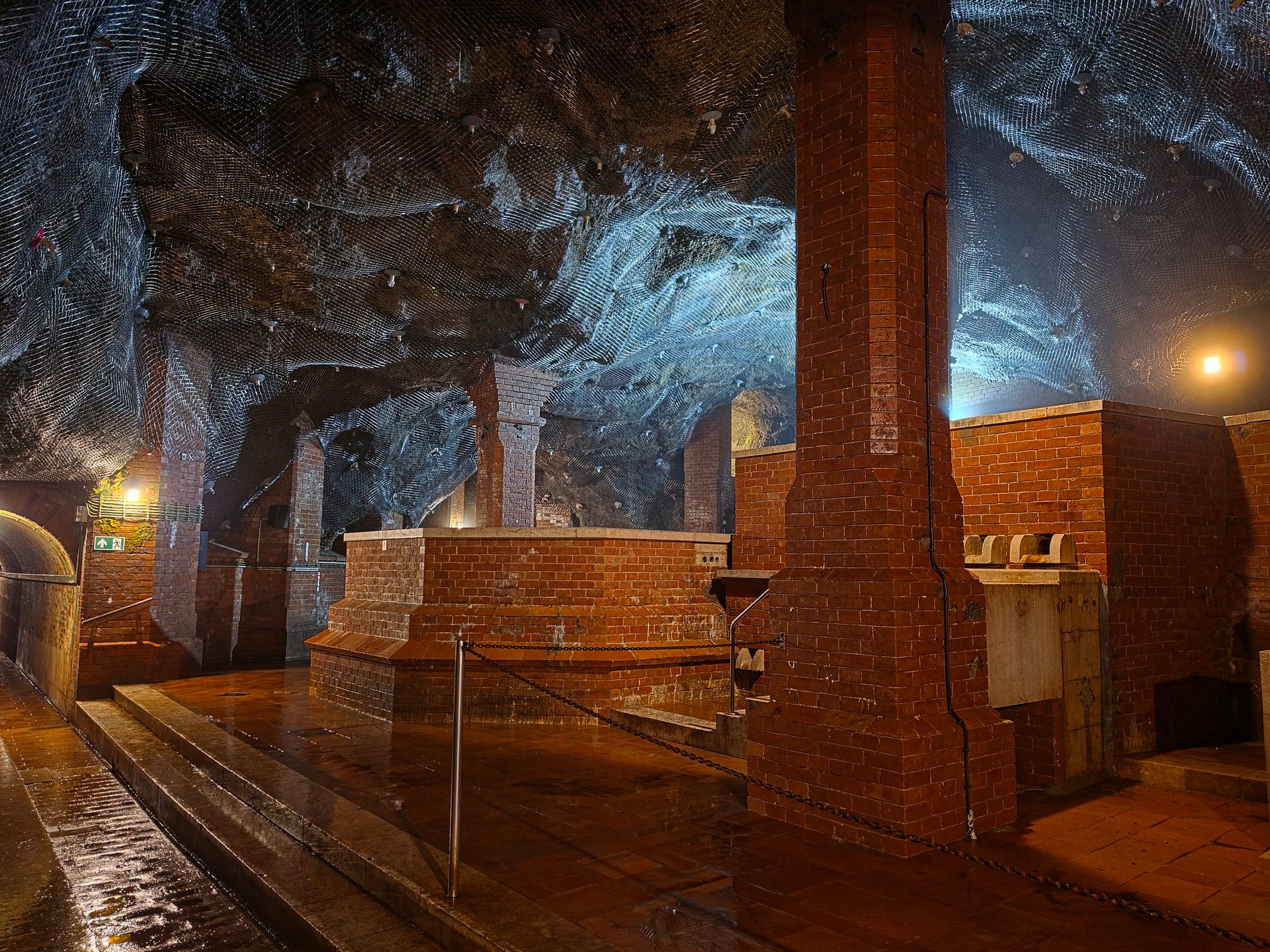 Underground chamber with robust brick pillars, walls, and platforms, a wet reflective floor, and a netted ceiling.