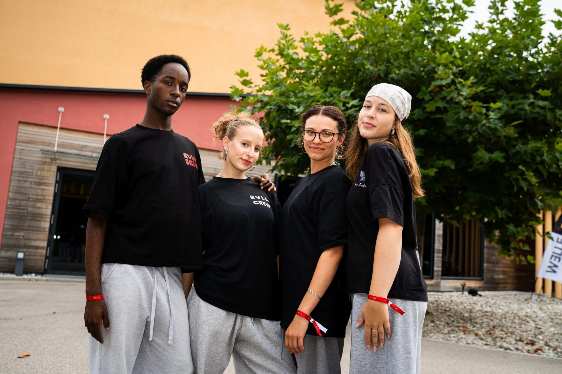 Four young people in black t-shirts and gray sweatpants with red wristbands pose outdoors.
