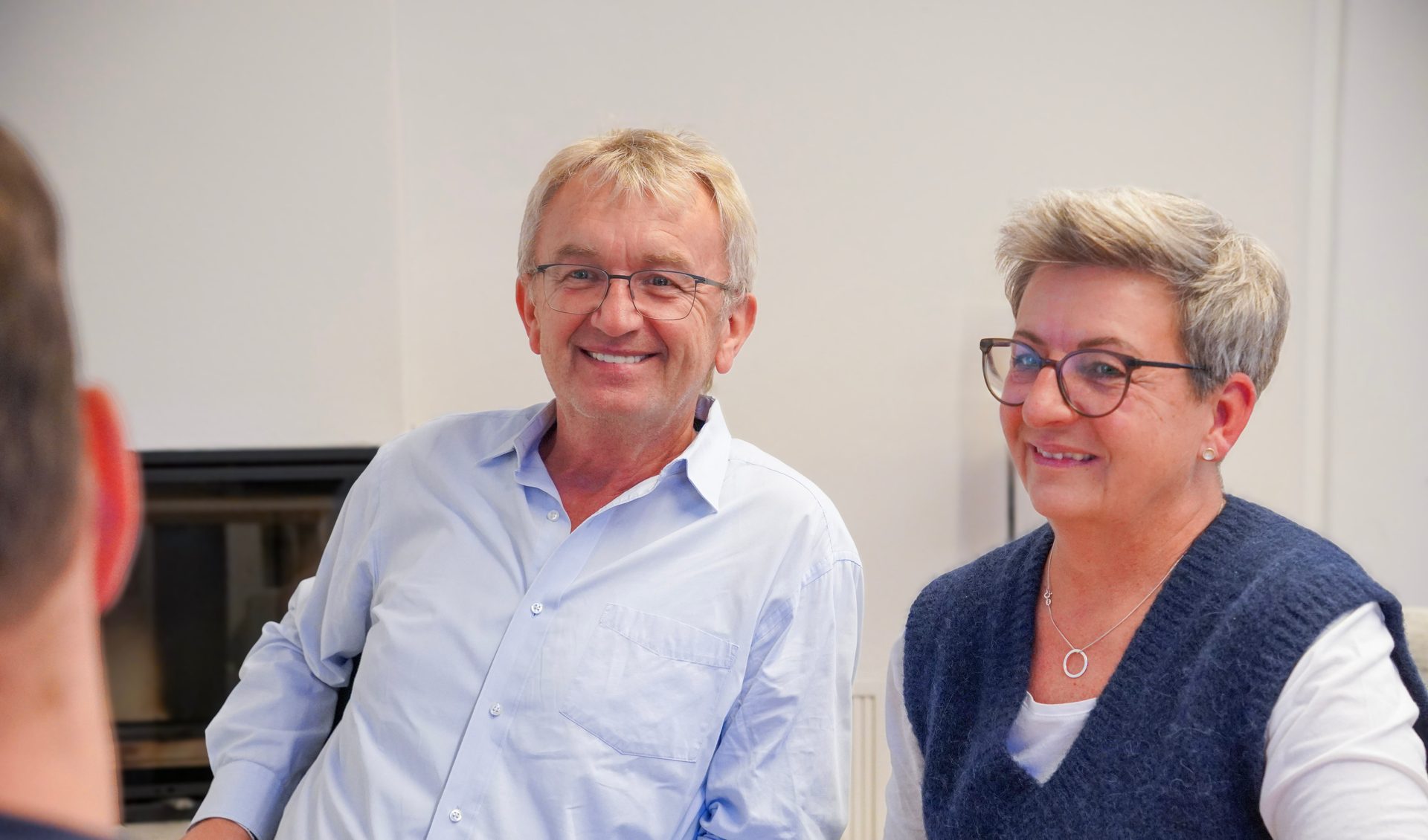 Two smiling people, a man and a woman in glasses, sit next to each other, looking forward.