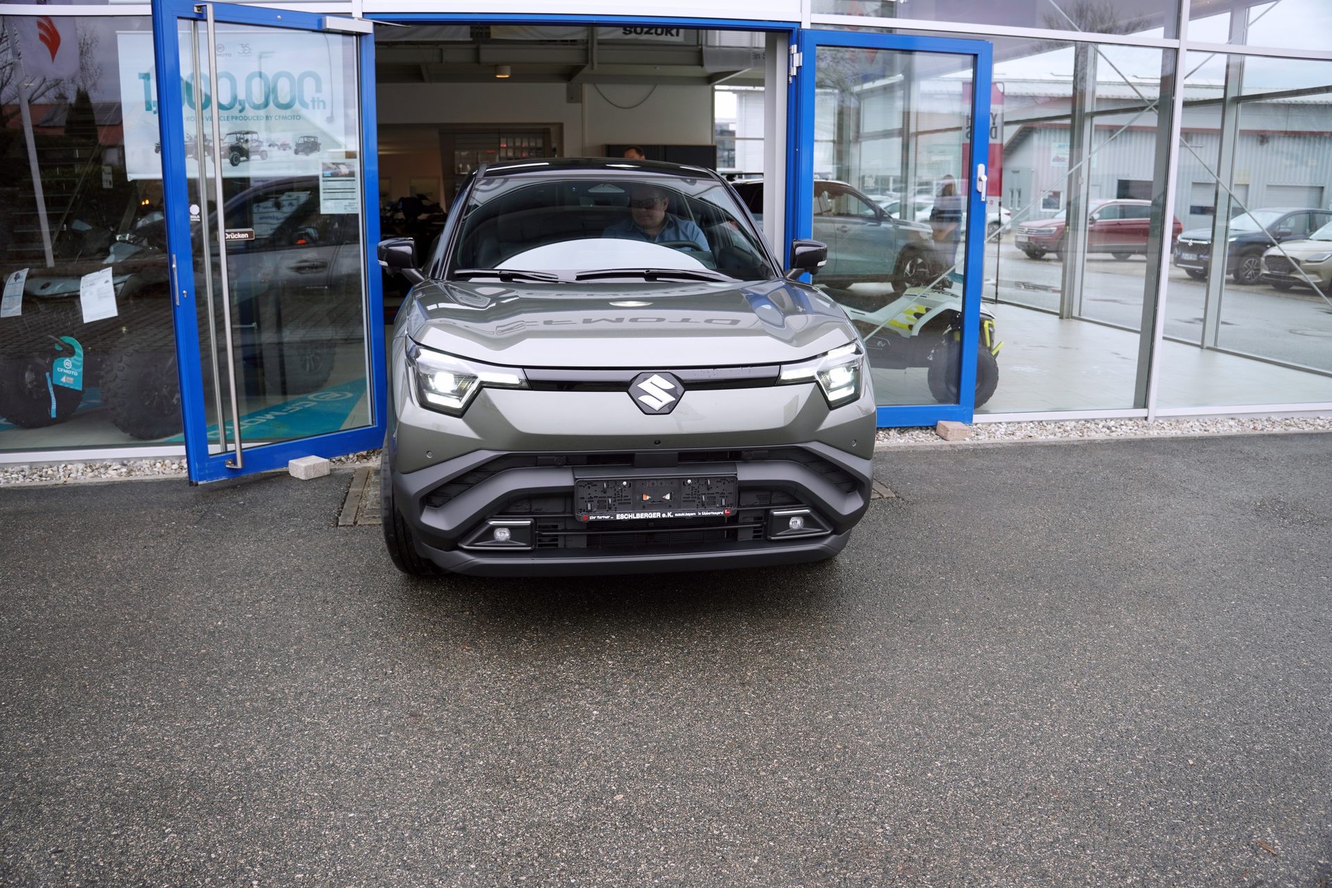 A grey SUV, possibly a Suzuki Fronx, is partially parked outside a car dealership showroom.