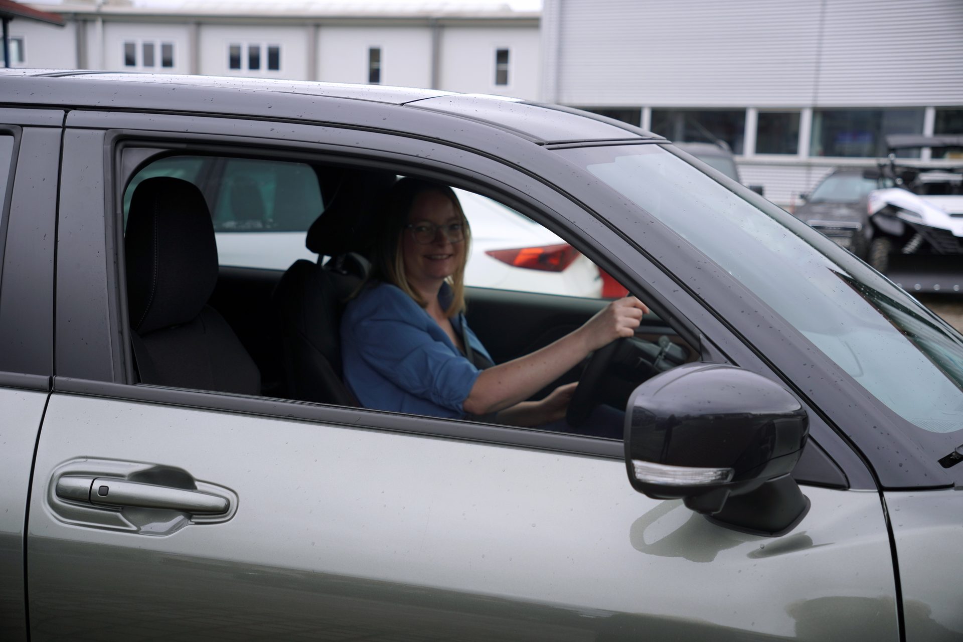 A smiling woman in a blue shirt and glasses is driving a gray car, looking directly at the camera.