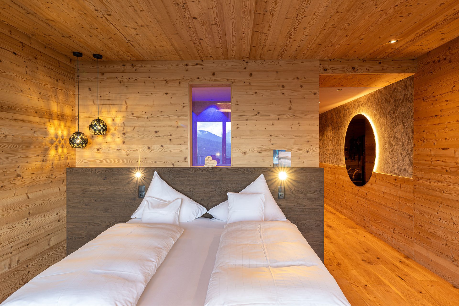 A cozy bedroom with a wooden interior, featuring a large bed with white bedding, modern lighting, and a mountain view.