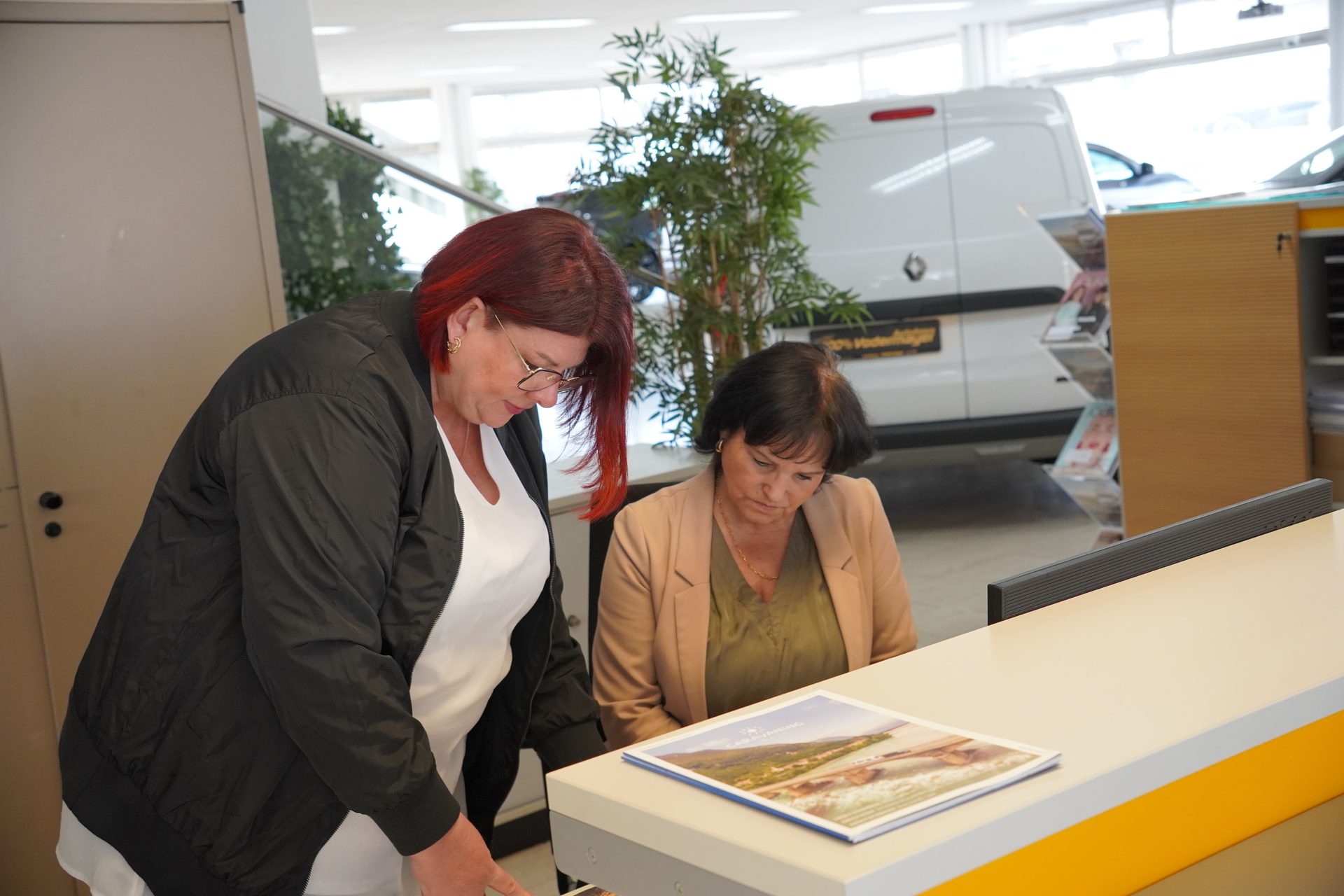 Two women, one with red hair, reviewing a document together on a counter in a car dealership.