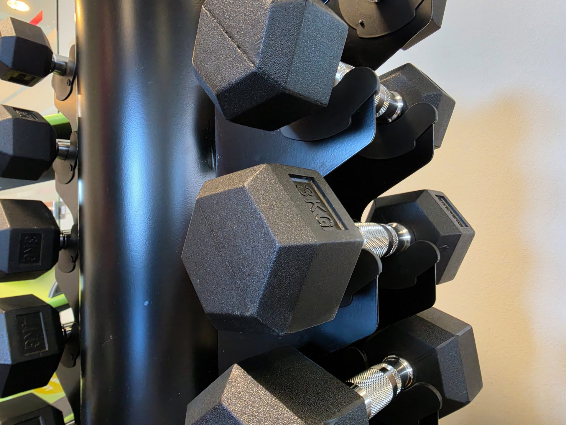 A close-up shot of black hexagonal dumbbells neatly arranged on a vertical rack in a gym, showing 3kg and 5kg weights.