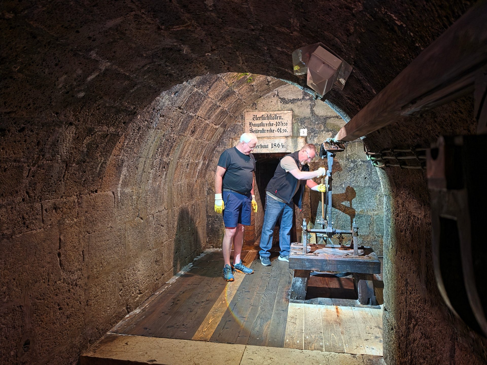Two men in an old stone tunnel, one operating historical machinery, the other observing.