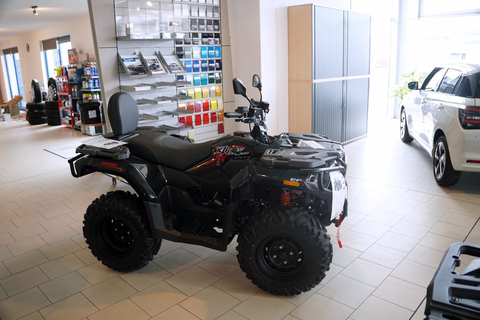 Black ATV (all-terrain vehicle) on display in a showroom with a white car and paint swatches.