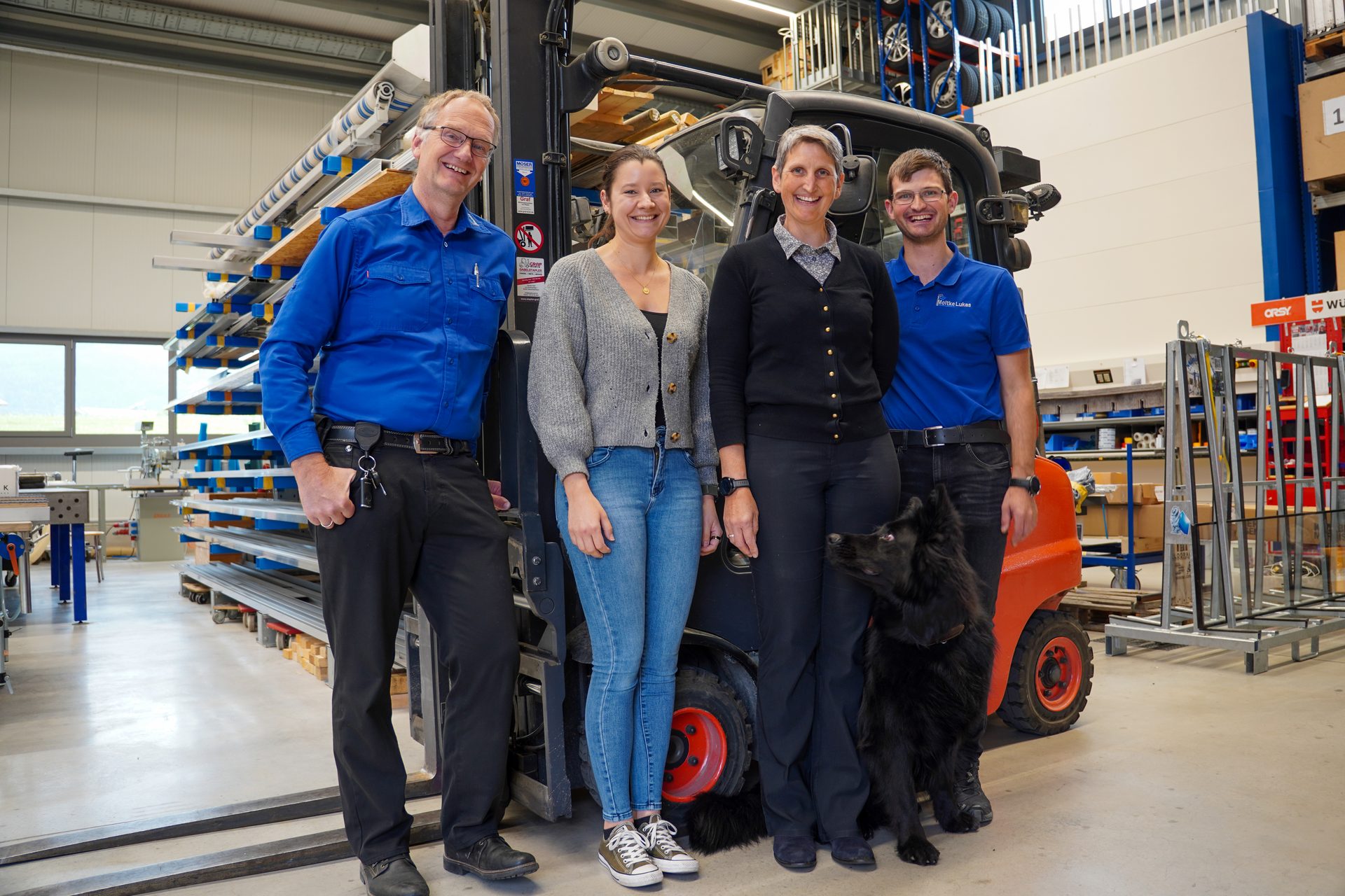 Four smiling people and a black dog stand in front of a forklift in a warehouse.