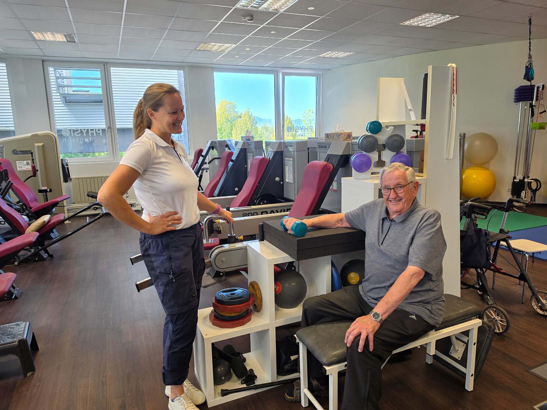 Physiotherapist assists an elderly man with dumbbell exercises in a modern gym.