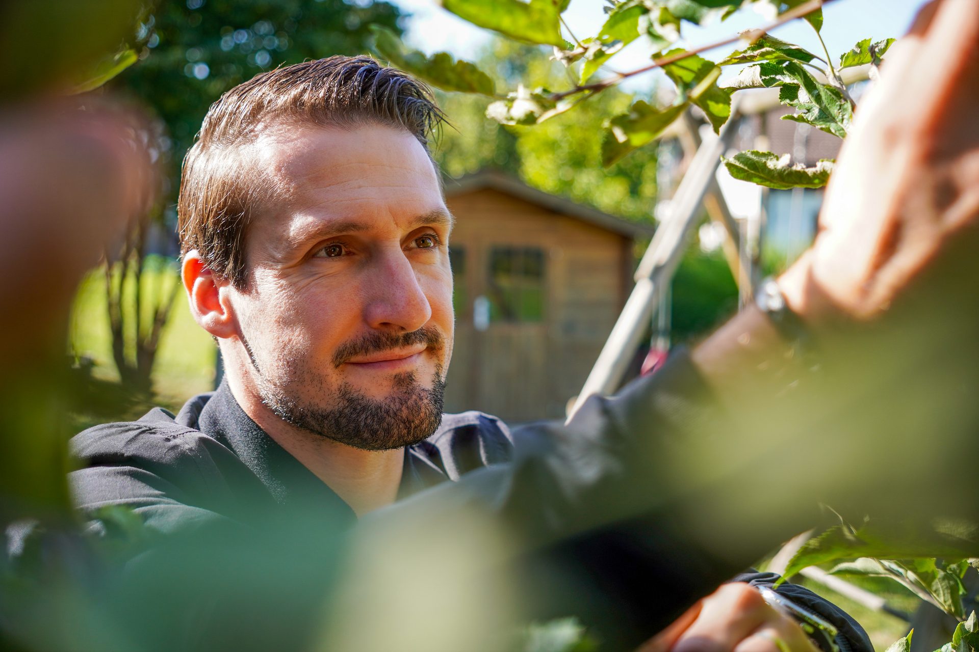 A man with a beard, framed by green leaves, is looking to the right with a slight smile.