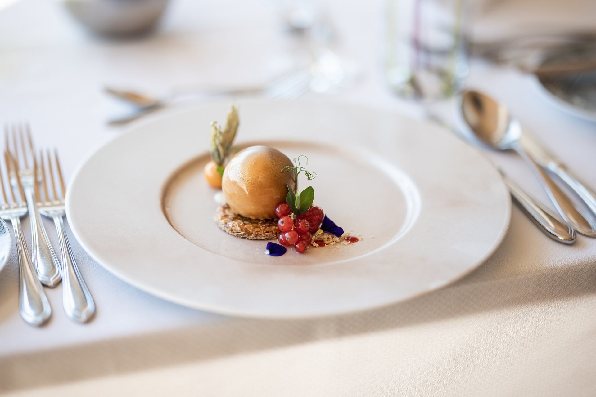 Elegant plated dessert with a spherical caramel mousse, red currants, and physalis on a white plate.