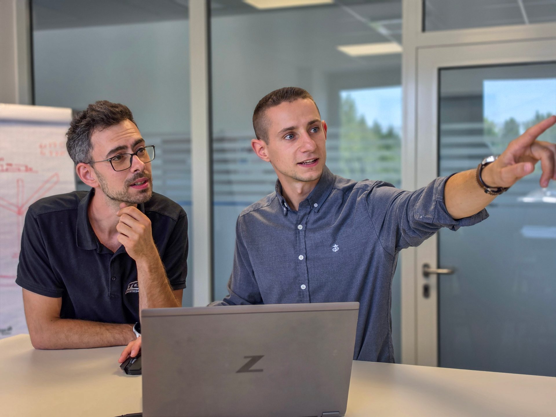 Two men in an office discussing, one pointing while explaining, the other listening intently.