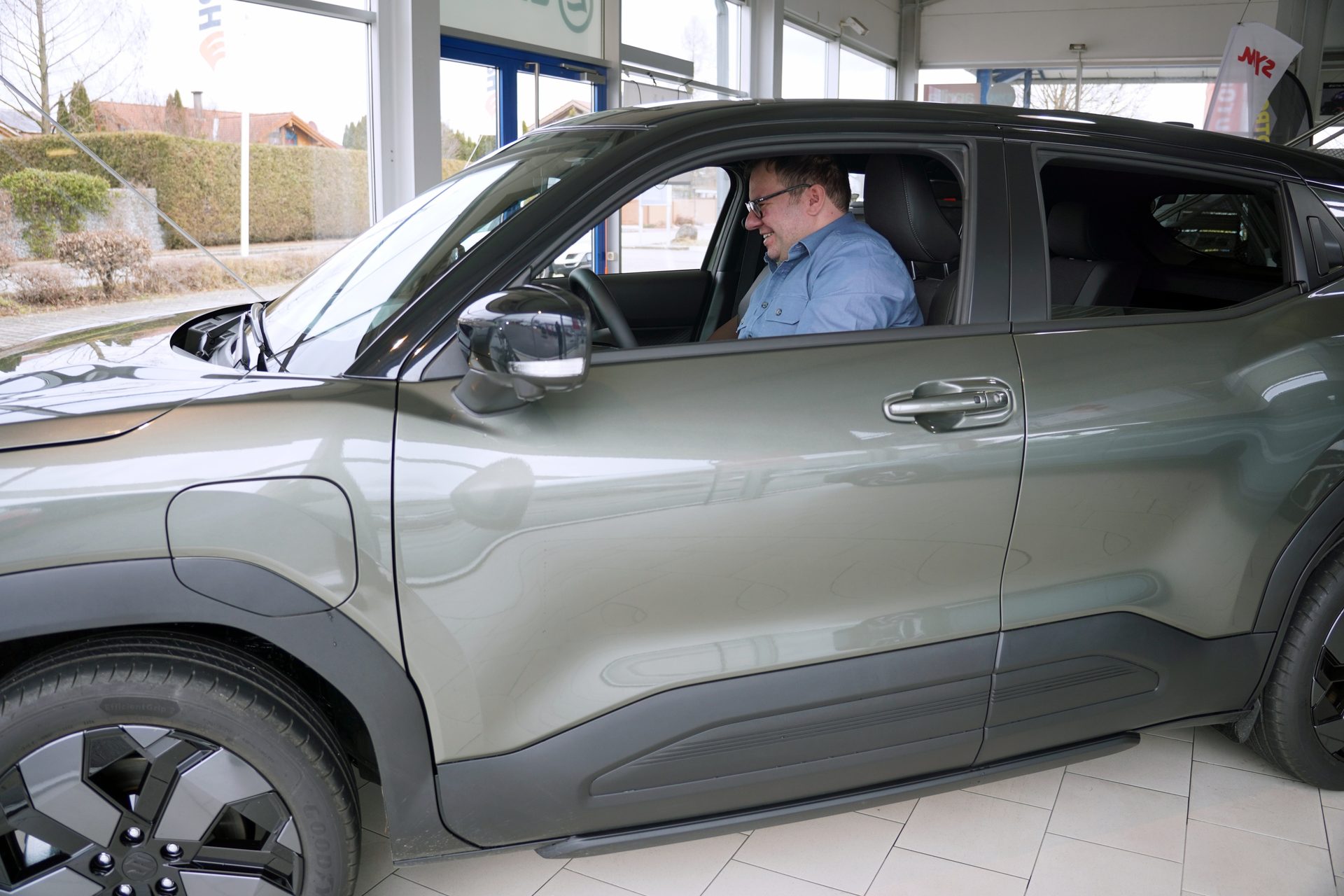 A man with glasses is laughing while sitting in the driver's seat of a new green car in a showroom.