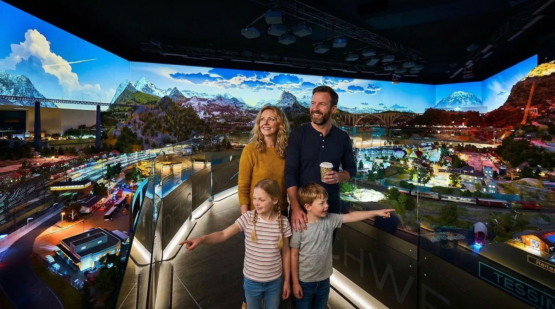 A family of four, two adults and two children, stands admiring a large, detailed miniature railway and landscape display.