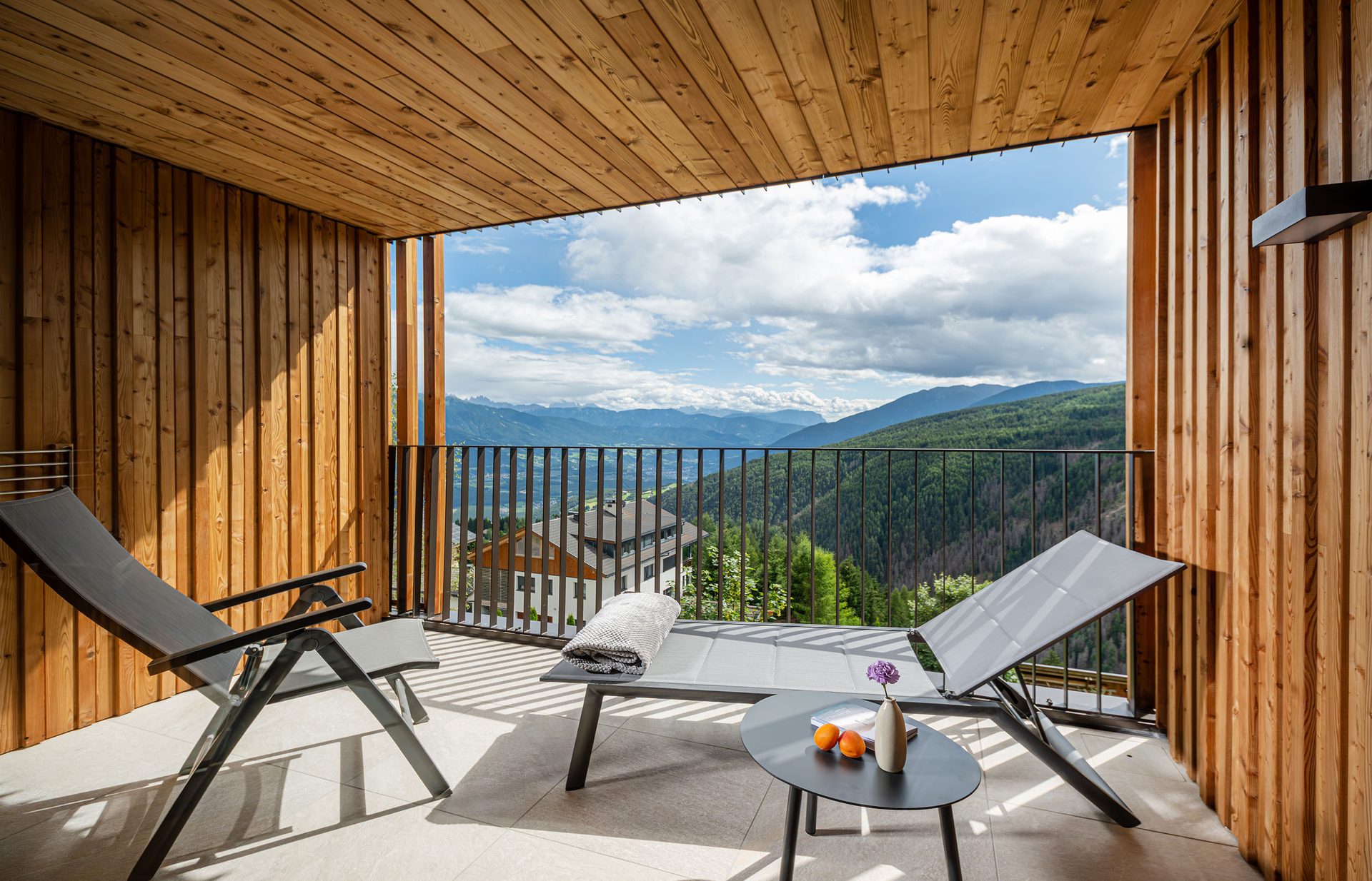 A wooden balcony with two lounge chairs and a small table, offering a panoramic view of mountains and a valley under a cloudy sky.