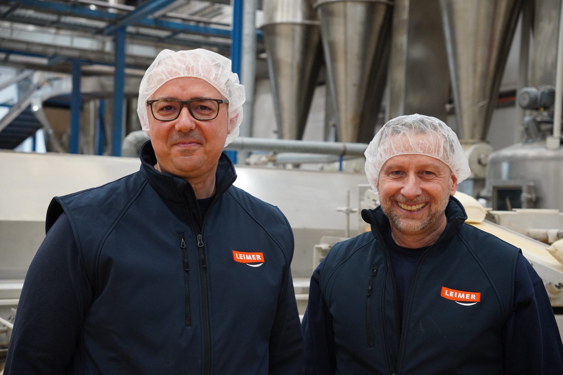 Two men in hairnets and 'LEIMER' vests smile in a factory setting.