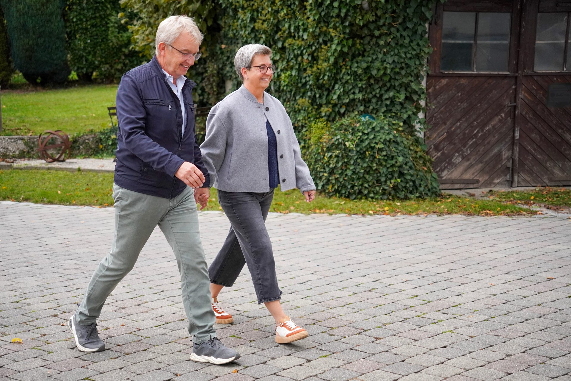 Two smiling people, a man and a woman, walk together on a paved path with greenery in the background.