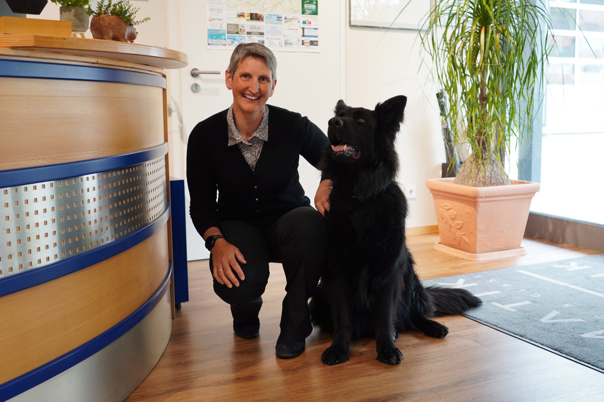 A woman kneels next to a large black dog, both looking at the camera and smiling in an indoor setting.
