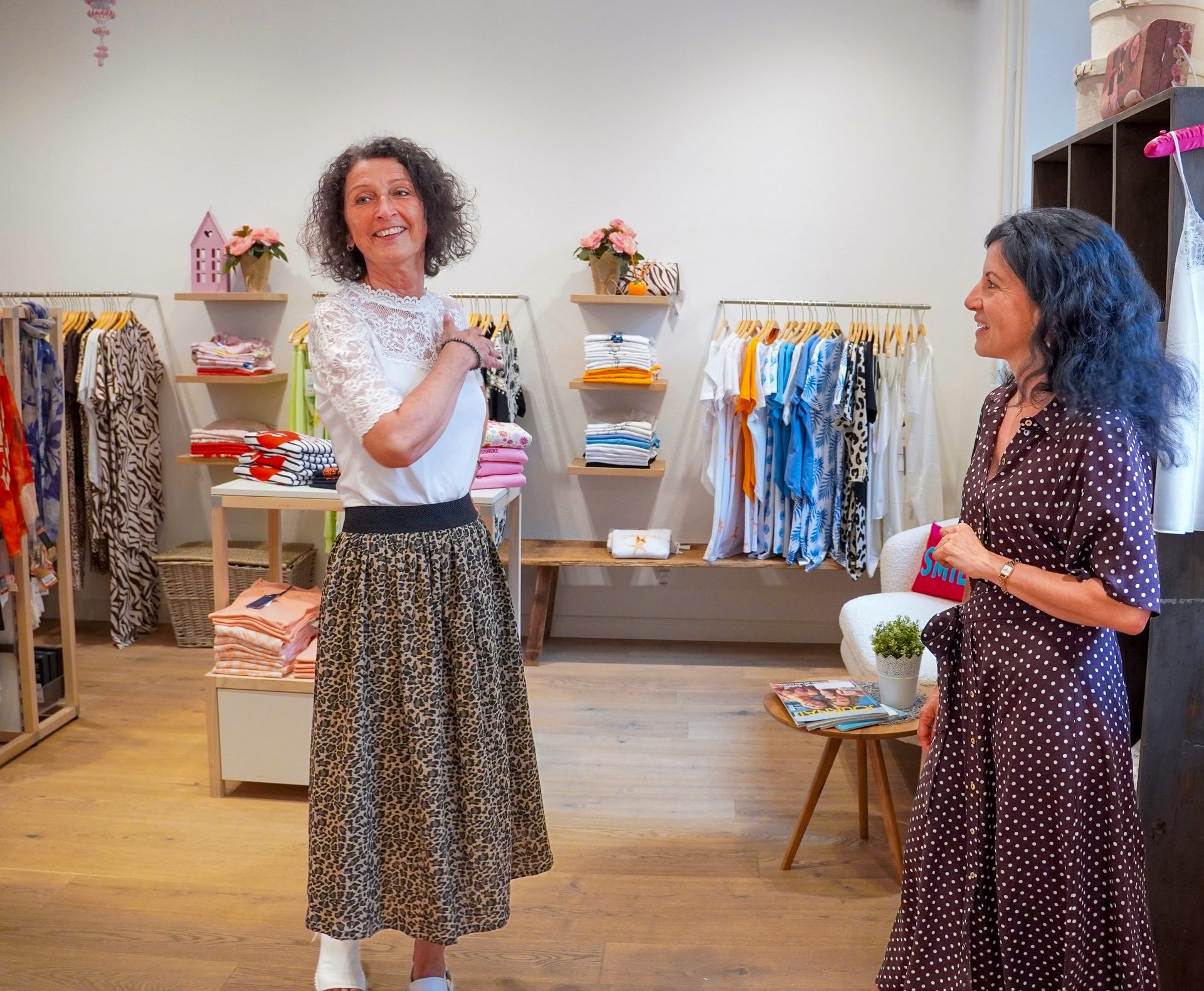 Two women chatting and smiling in a clothing boutique with racks of clothes.