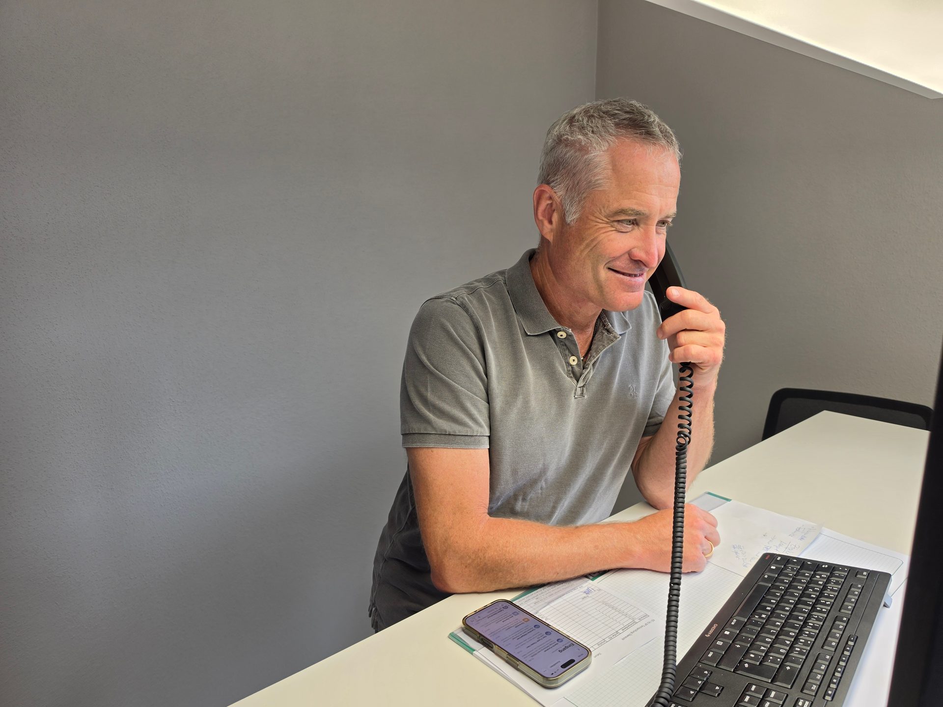 A smiling man in a grey polo shirt talks on a landline phone at a white desk with a smartphone and keyboard.