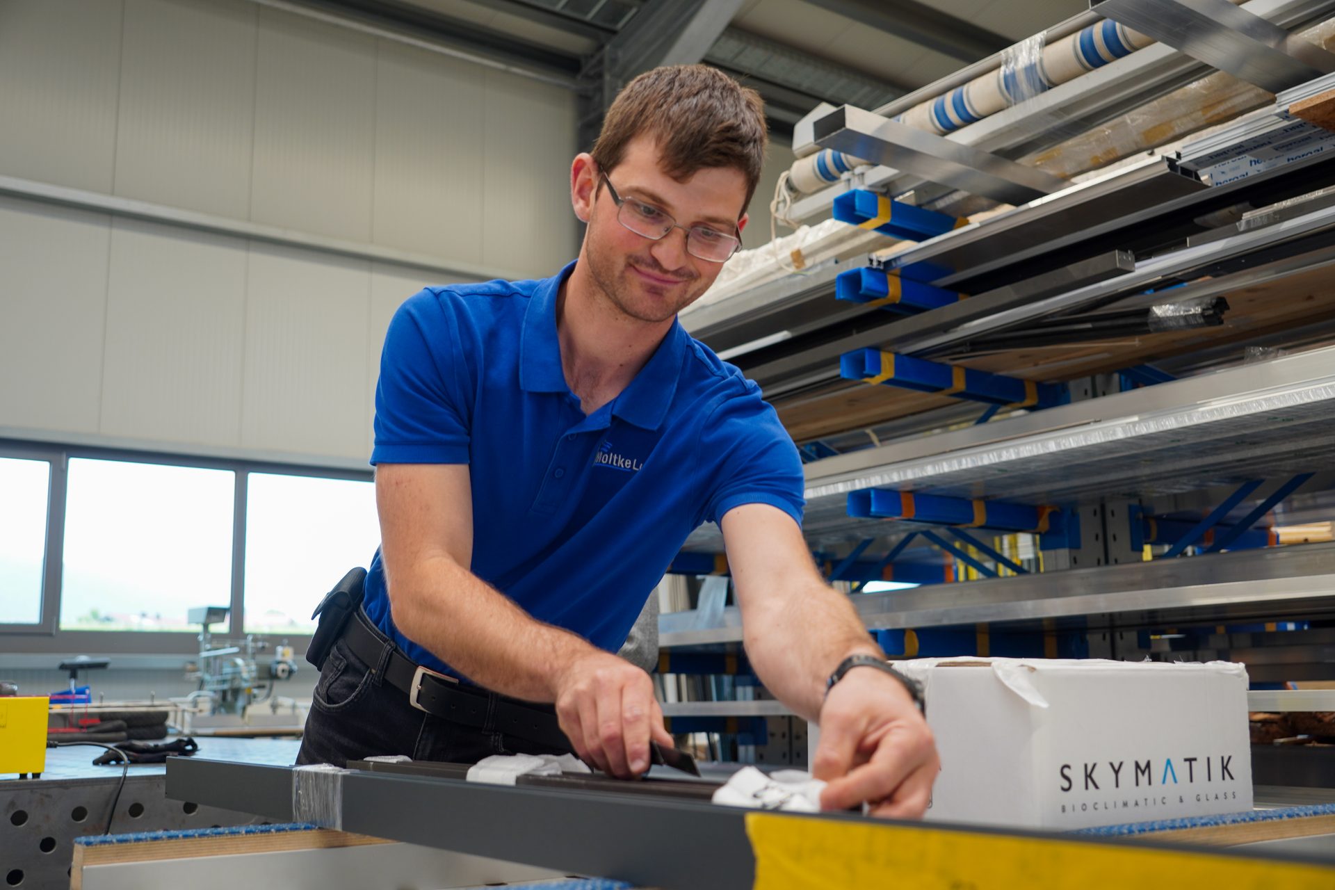 A man in a blue shirt and glasses working on metal profiles in a workshop.