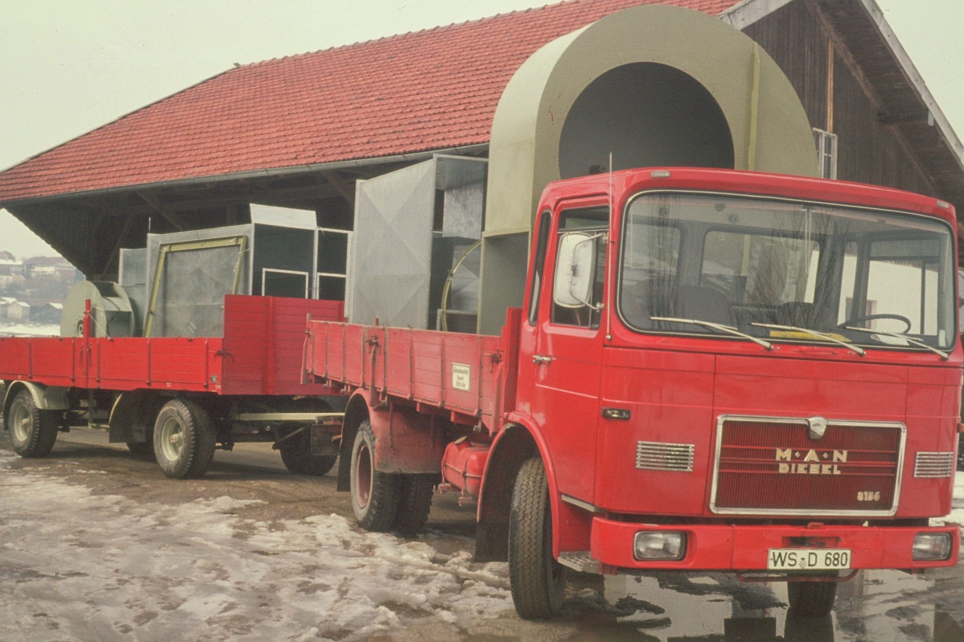 A red MAN Diesel truck and trailer loaded with large industrial ventilation units, parked on snowy ground.