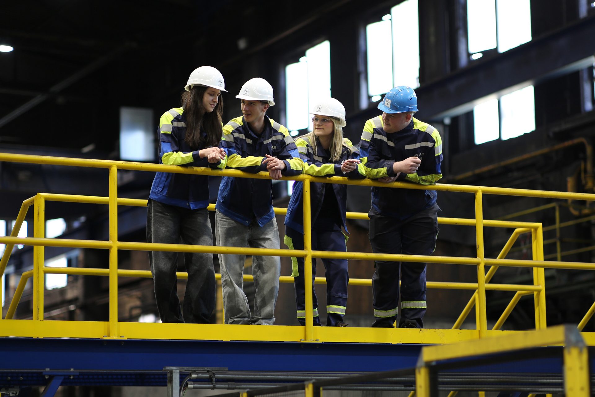 Four young workers in hard hats and safety jackets stand on a yellow platform in an industrial setting.