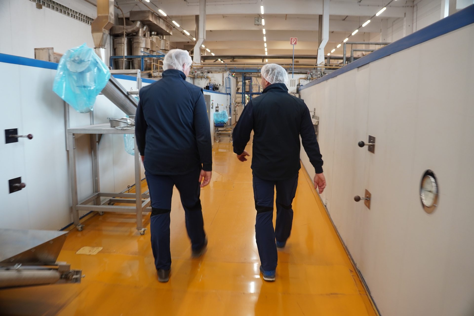 Two men in hairnets and work attire walk through a clean food processing facility with yellow floors.