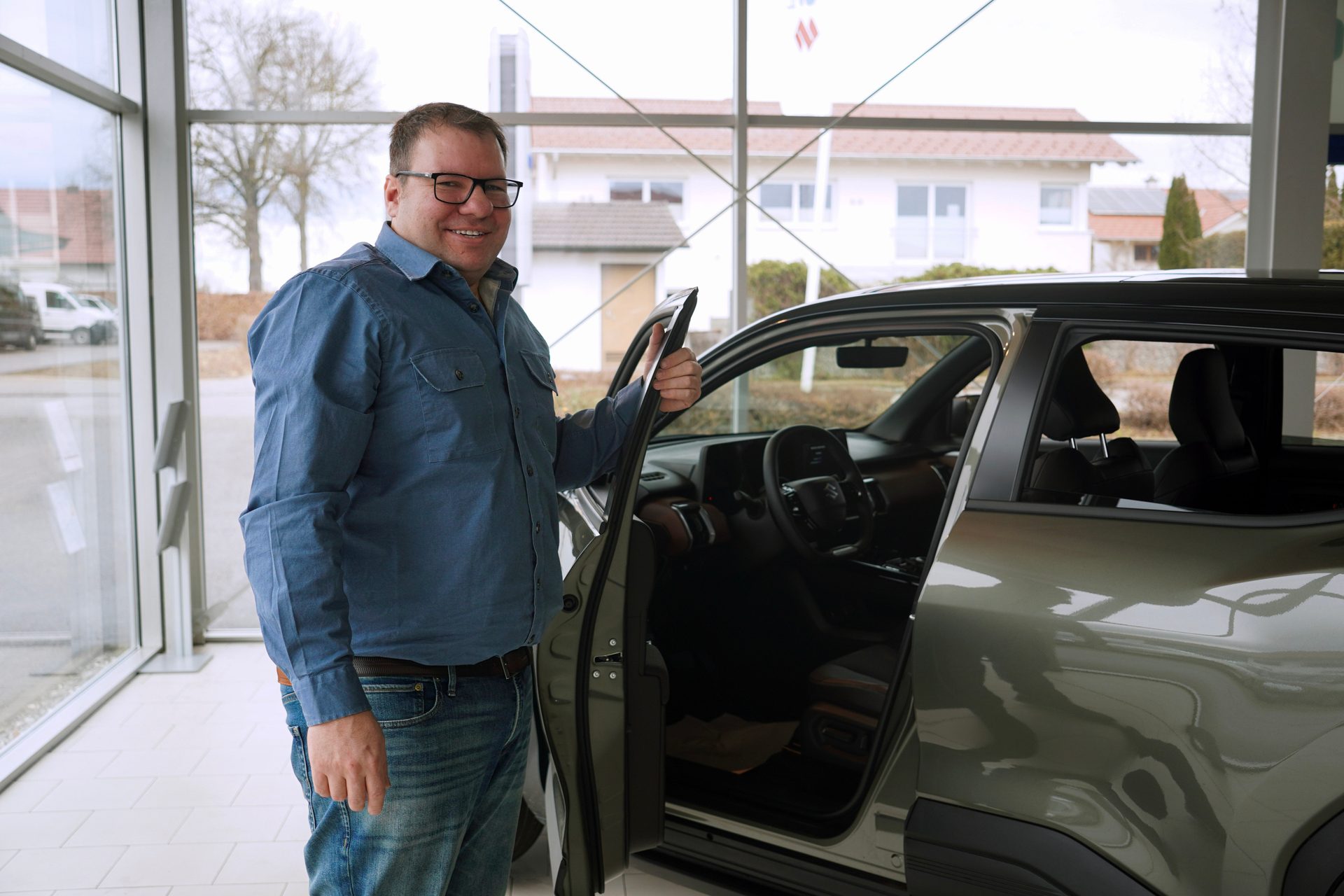 Man in blue shirt and glasses smiling next to an open car door in a dealership.