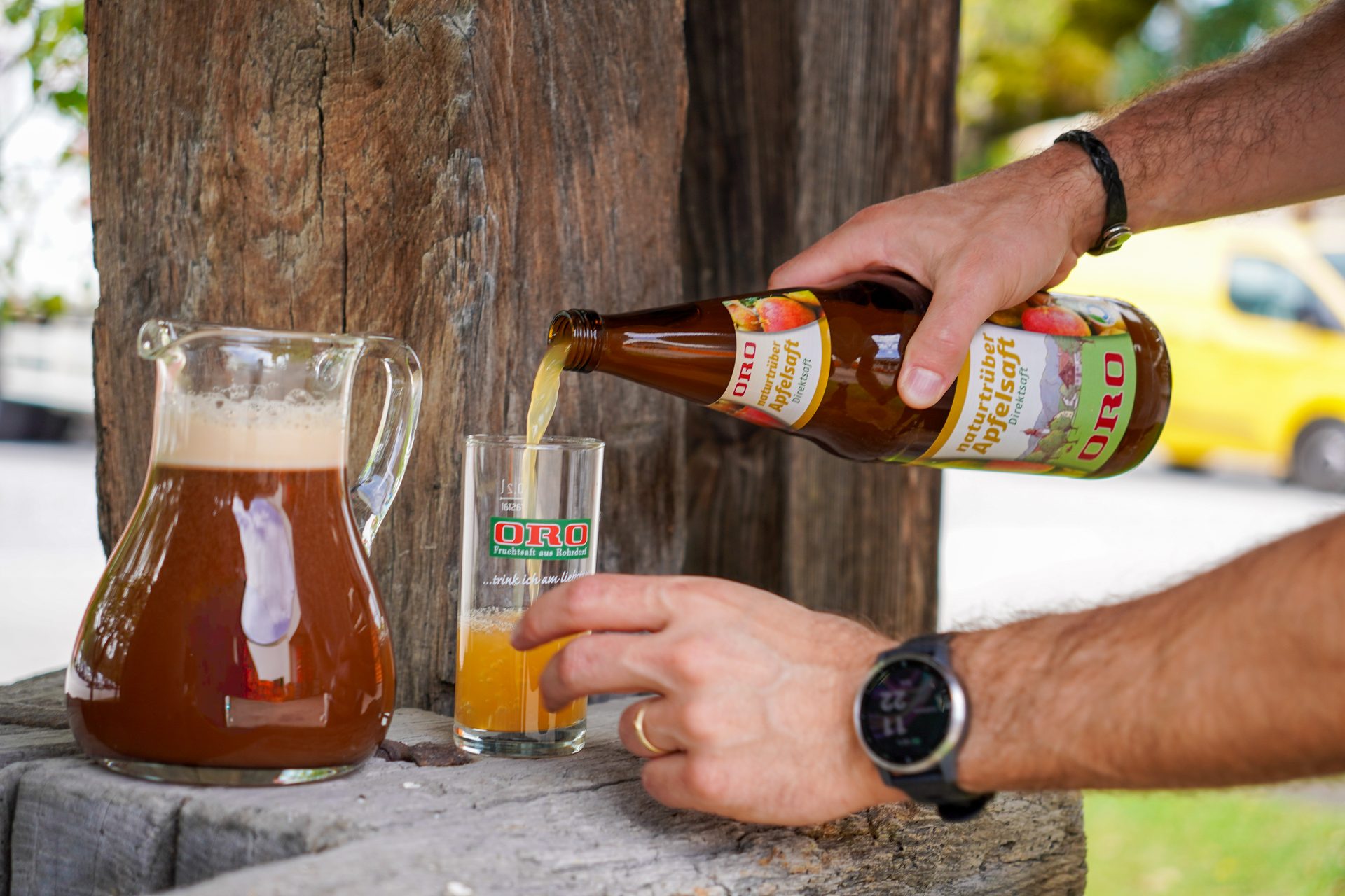 A person pours ORO apple juice into a glass from a bottle, with a jug on a rustic wooden table.