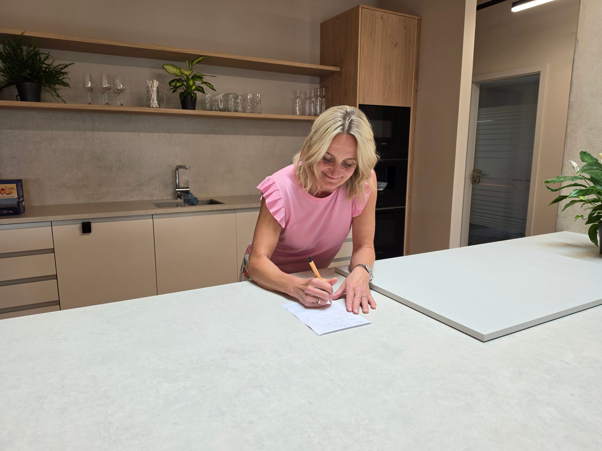 Blonde woman in a pink top writing on a piece of paper at a modern kitchen island.