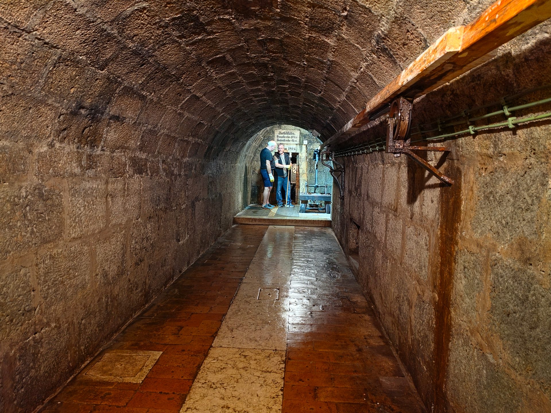 Two people stand at the end of a long, arched stone tunnel with an old wooden beam and pipes on the right.