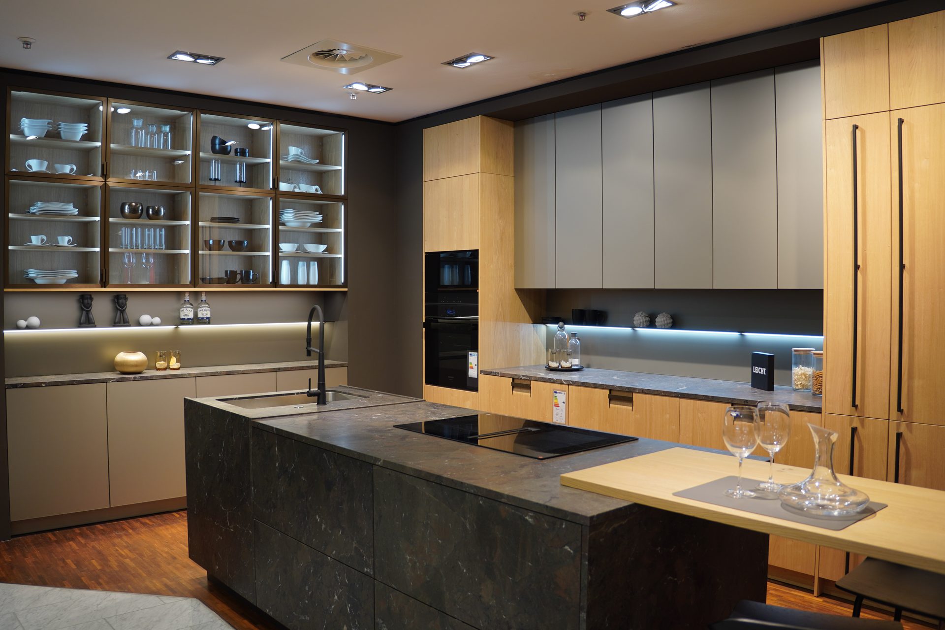 A modern kitchen with wooden and grey cabinets, a dark island, glass displays, and integrated appliances.