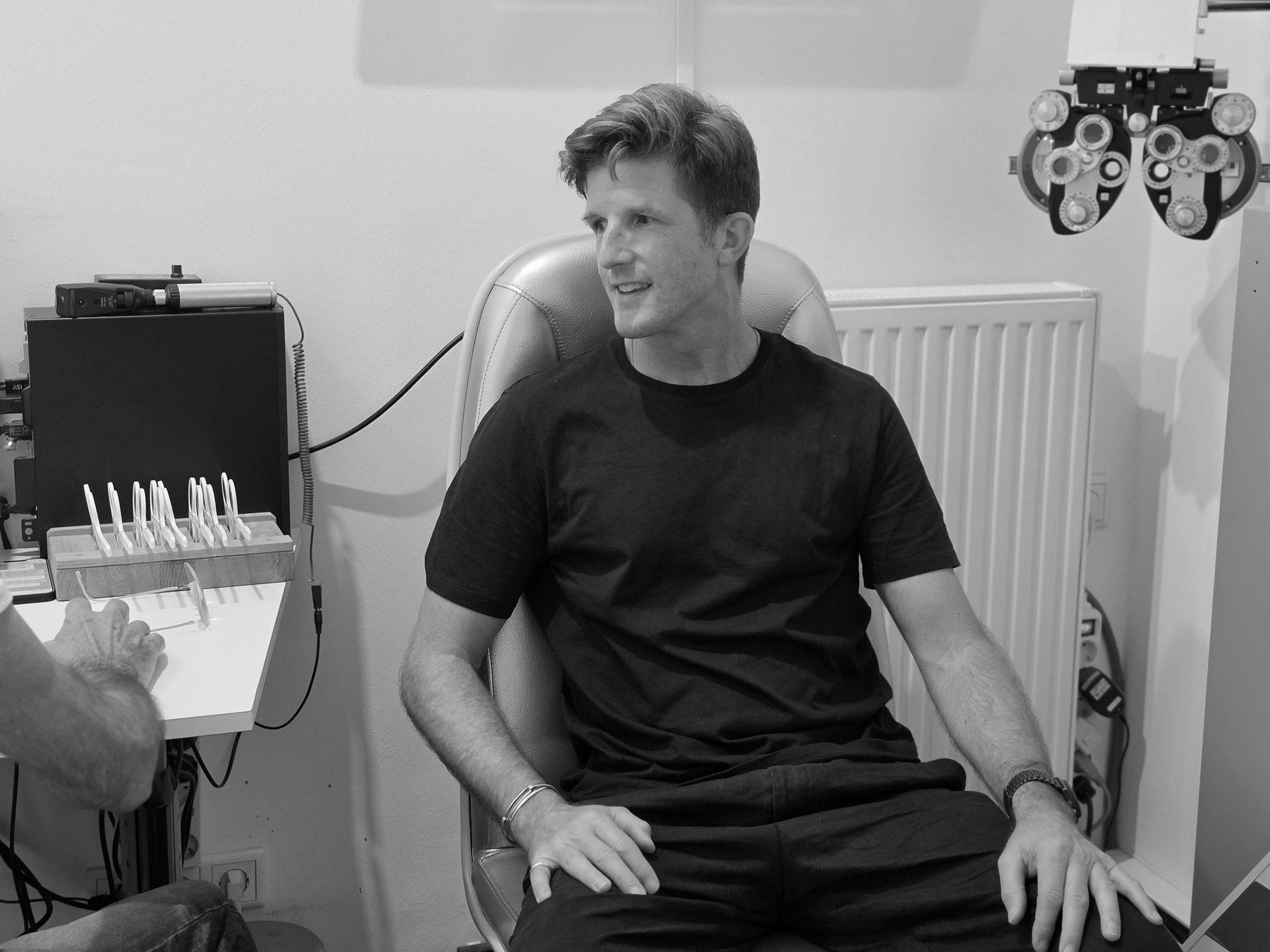 Man in black t-shirt sitting for an eye exam, with optometry equipment and glasses visible.