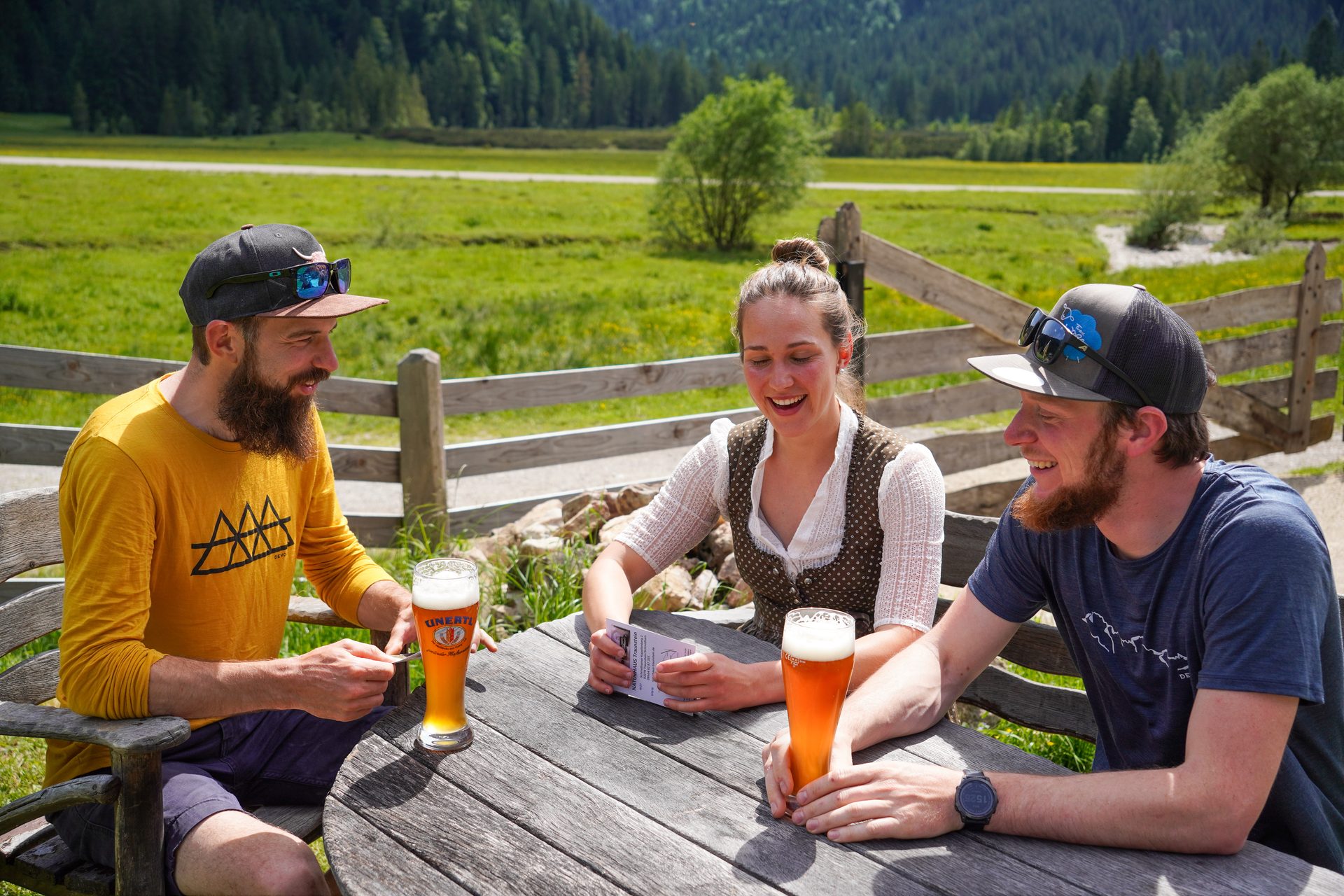 Three friends laugh over beers at a rustic outdoor table with a scenic mountain and meadow backdrop.