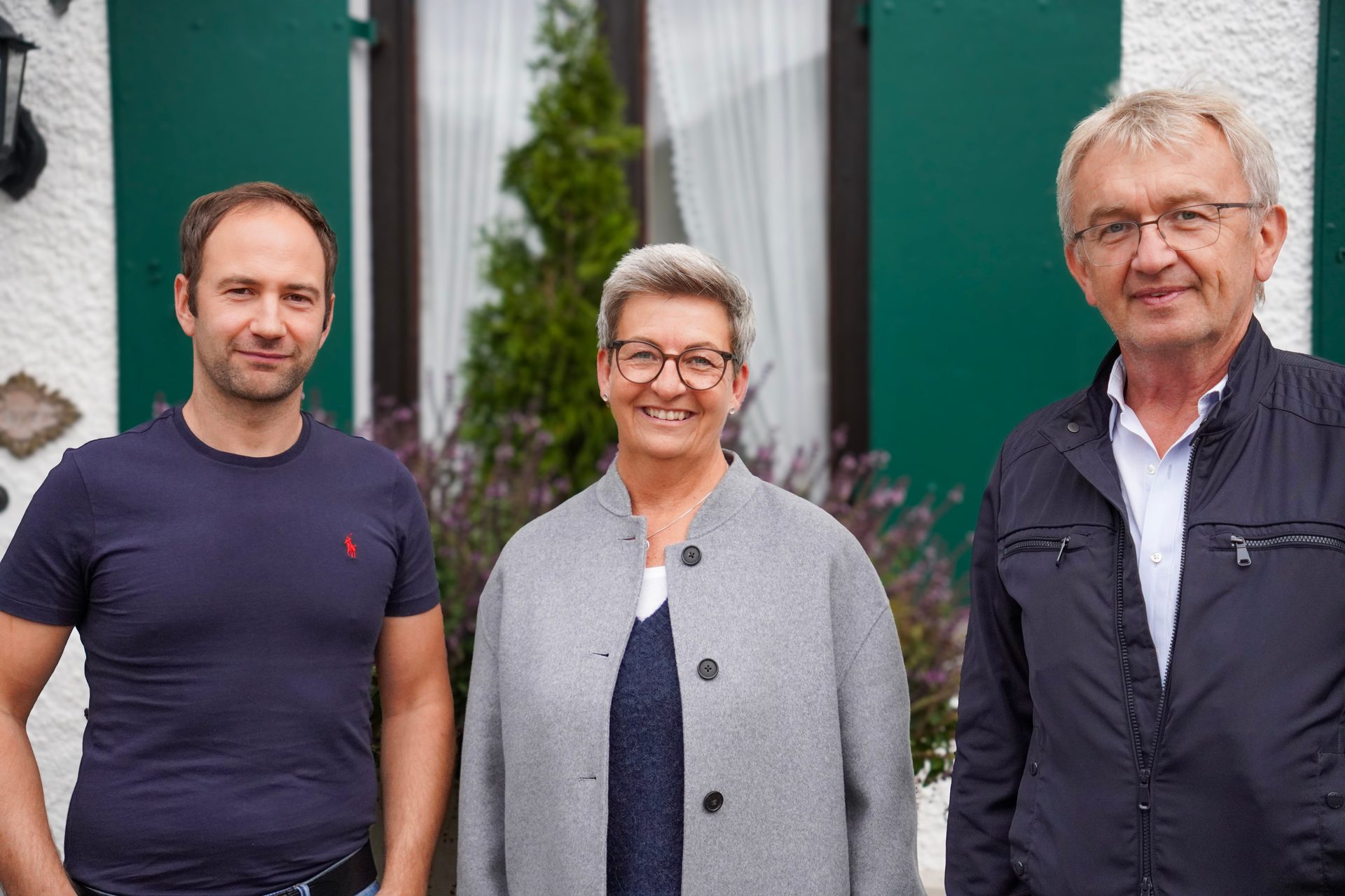 Three smiling people, two men and one woman, stand side-by-side outdoors against a green building.