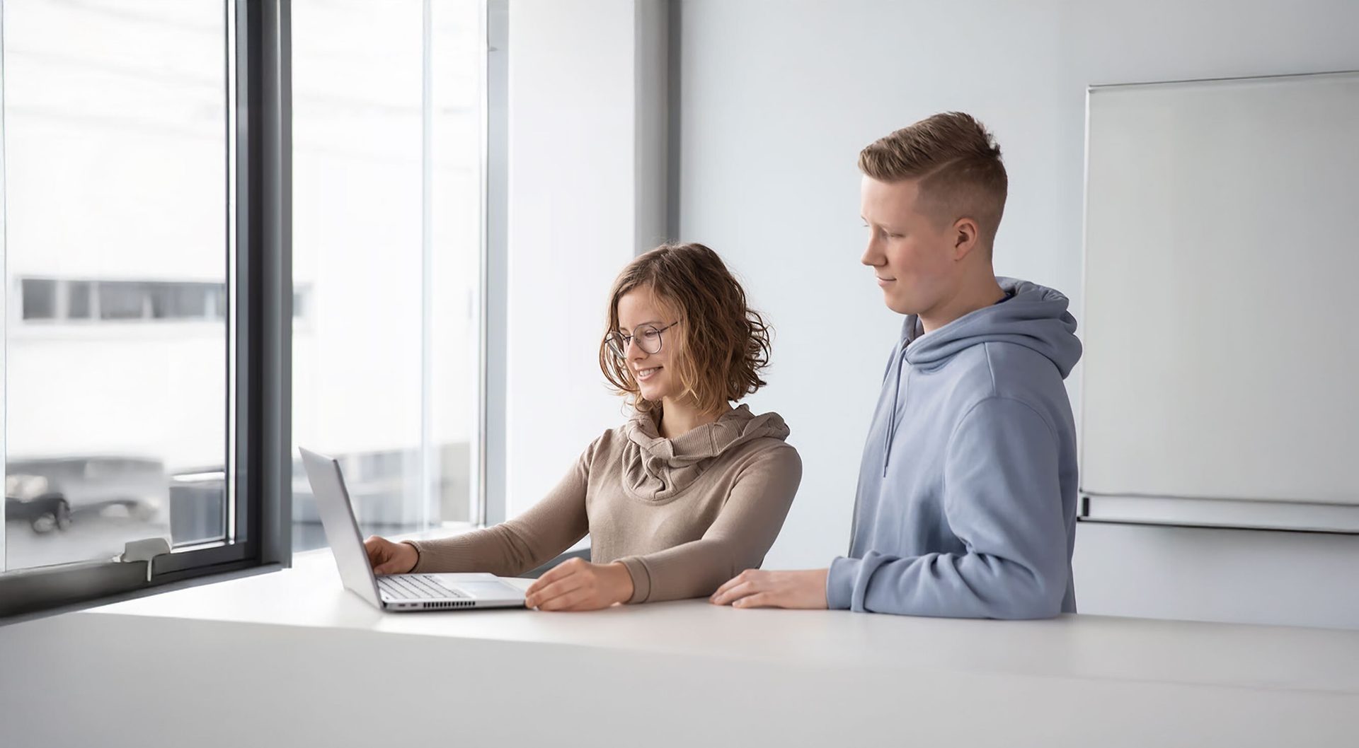 Young woman types on a laptop, while a man watches, by a bright window.