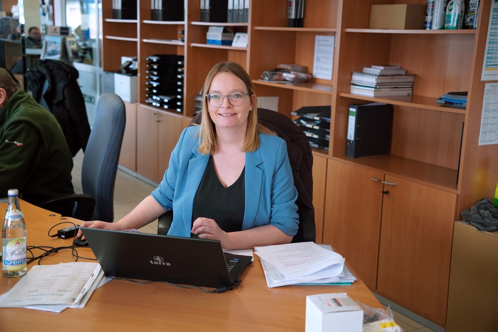 A smiling woman in a blue blazer works on a laptop at an office desk, with shelves in the background.