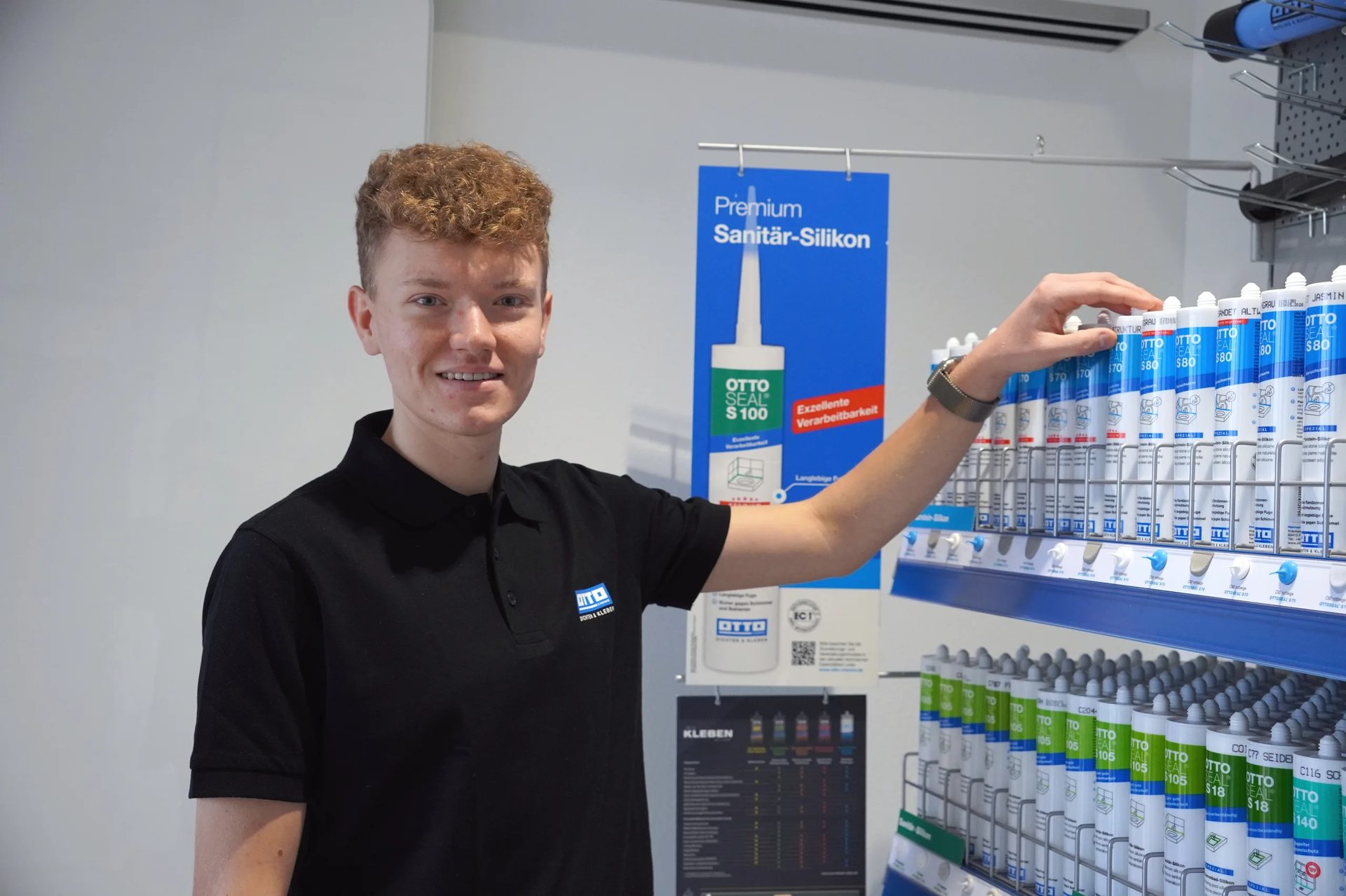 A young man in a black polo shirt smiles while touching sealant products on a store shelf, with a sign for "Premium Sanitär-Silikon" in the background.