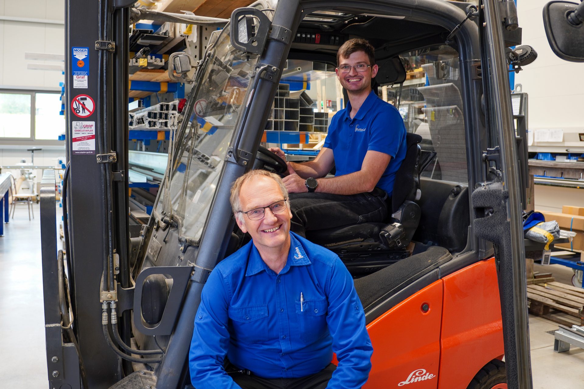 Two smiling men in blue shirts, one in a forklift and one standing, in a warehouse setting.