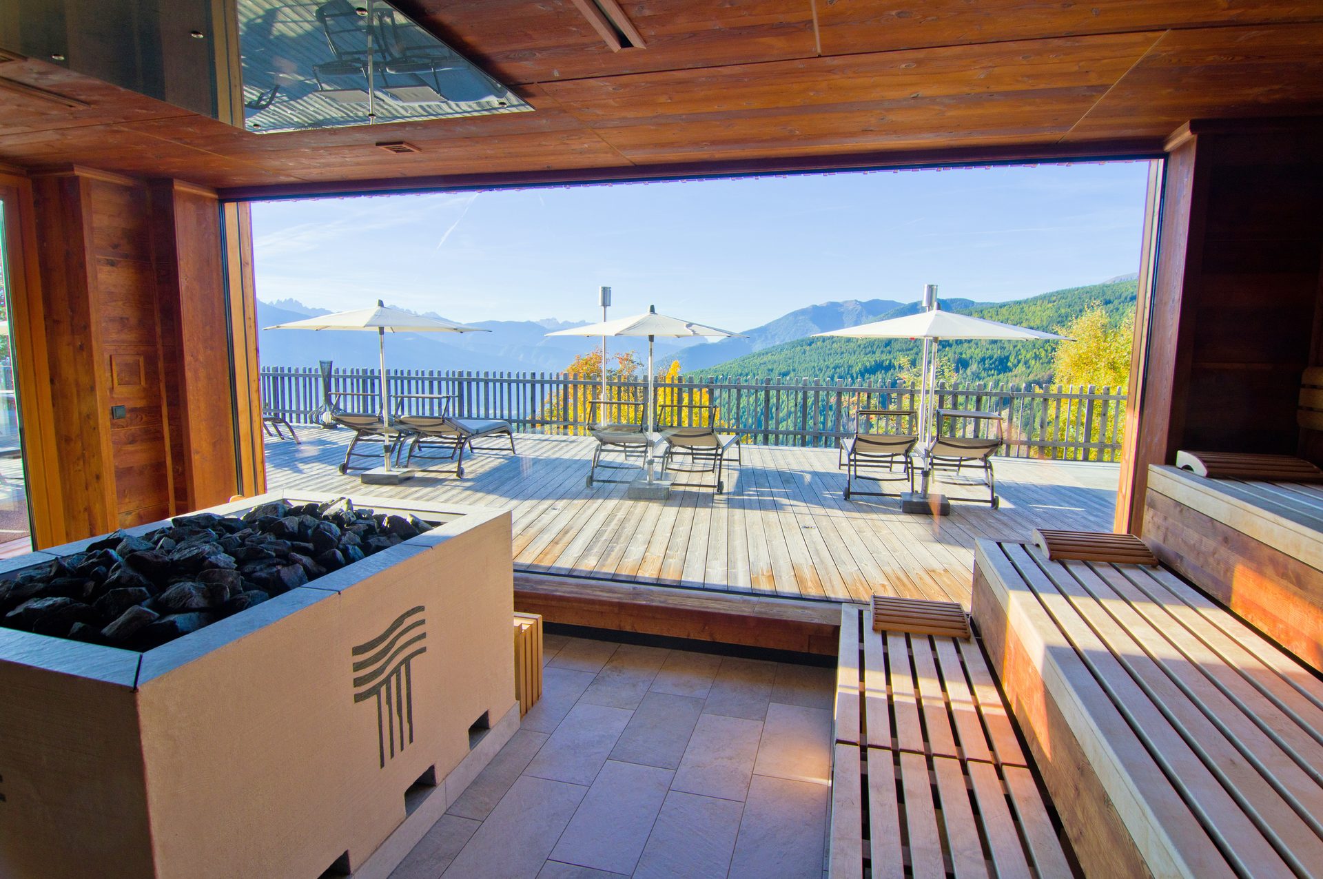 Luxurious indoor spa with wooden benches and a stone heater, overlooking an outdoor deck with mountain views.