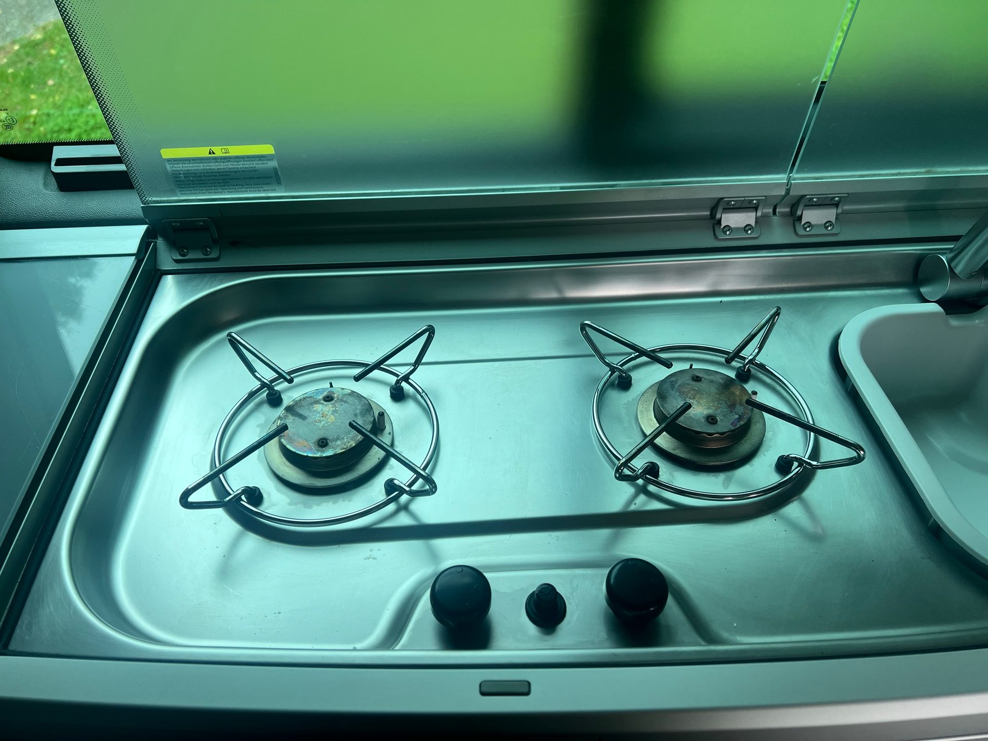 Two-burner camper stove and sink with open glass lid, showing used burners and control knobs.