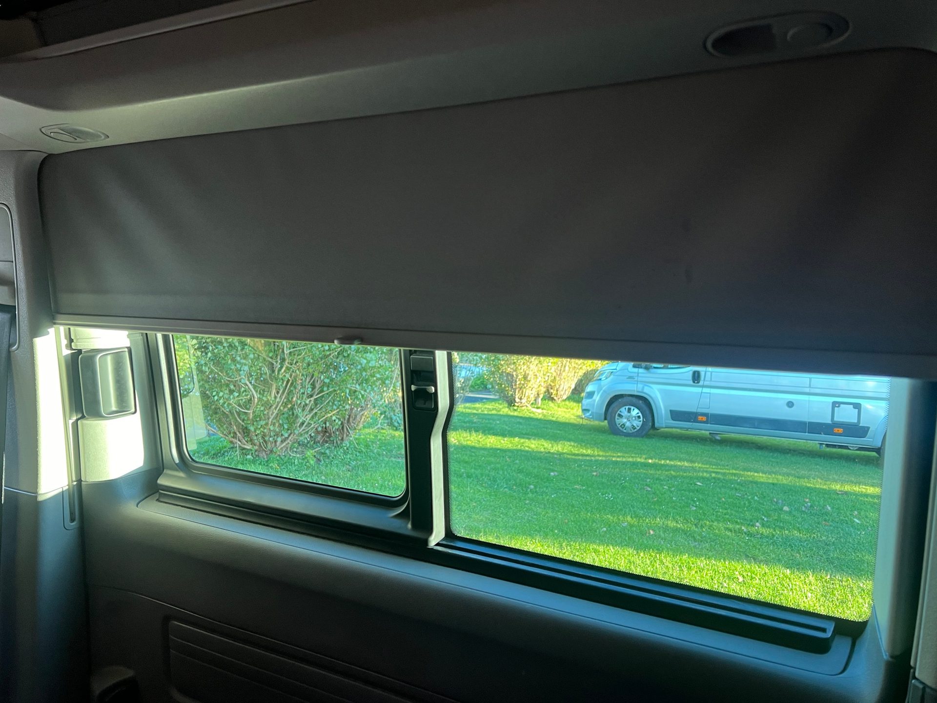 View from vehicle interior showing a partially lowered window blind, green grass, and a camper van outside.