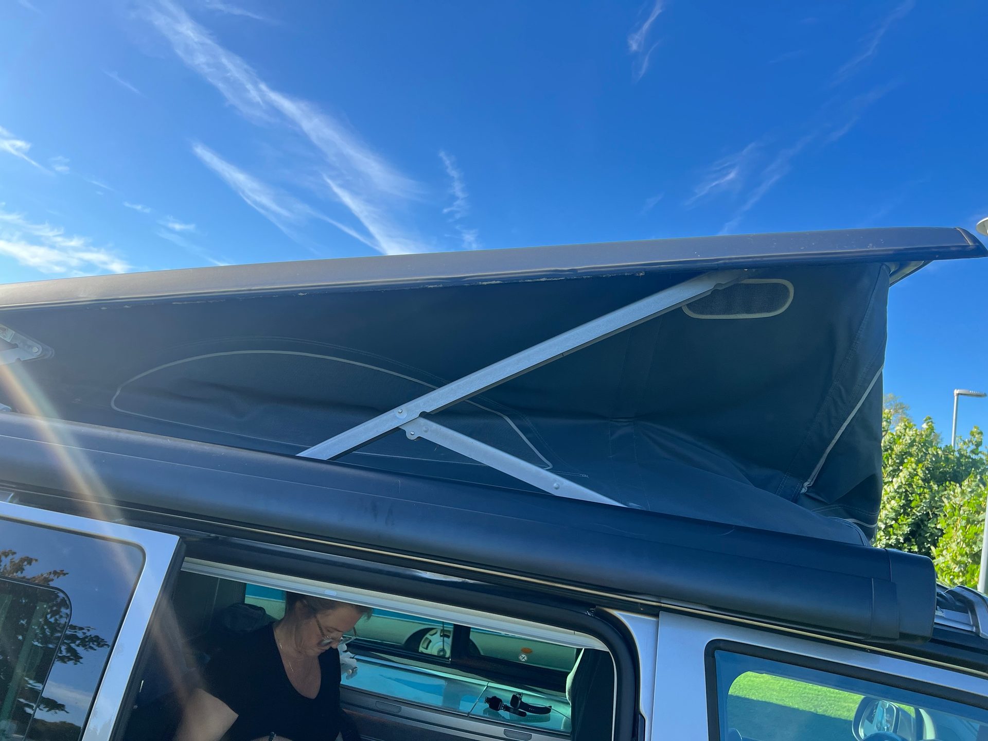 A woman sits inside a silver pop-top camper van, with its canvas roof extended under a blue sky.