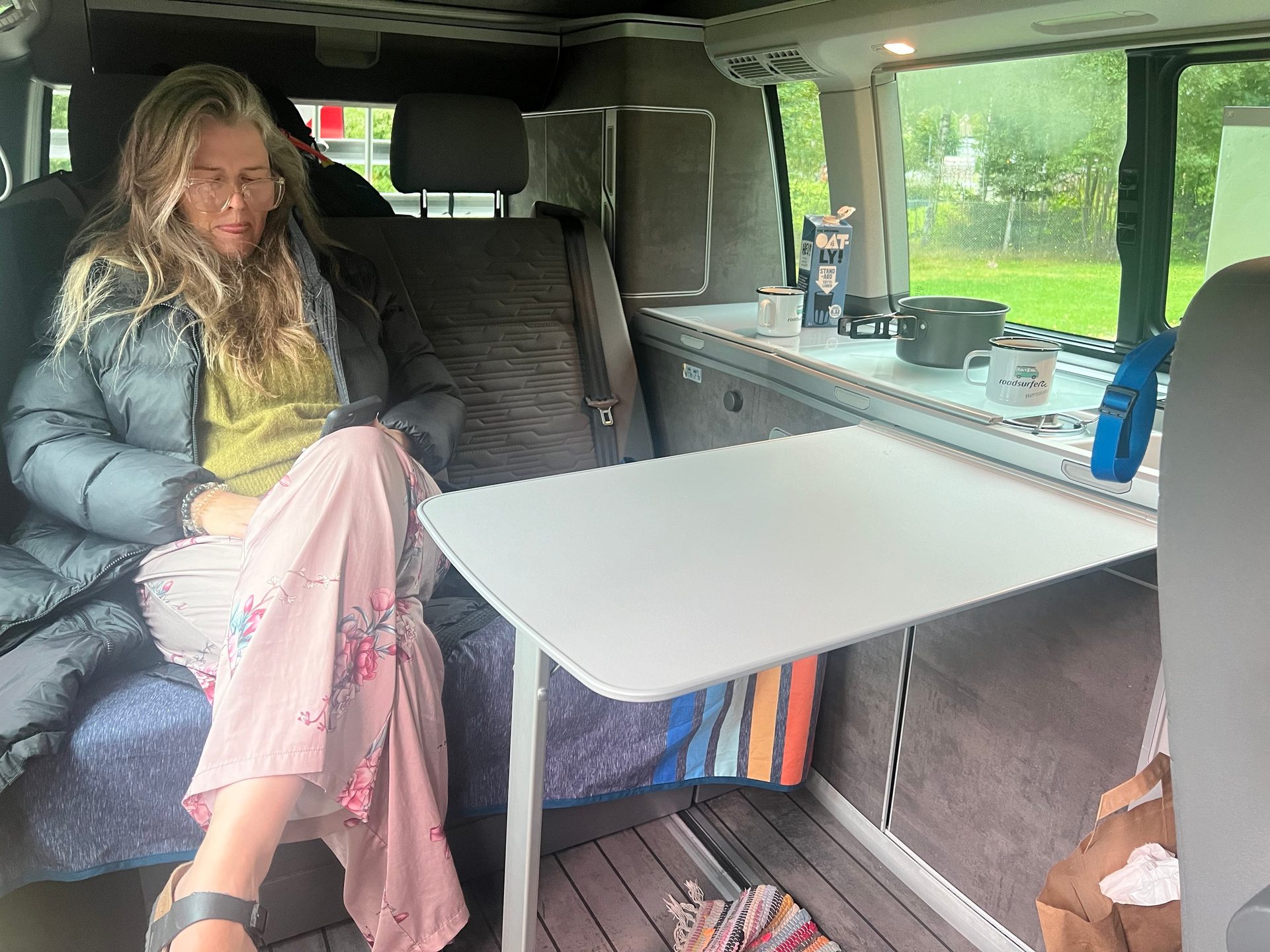 A woman in glasses, a puffer jacket, and floral pants, relaxes inside a camper van kitchen area.