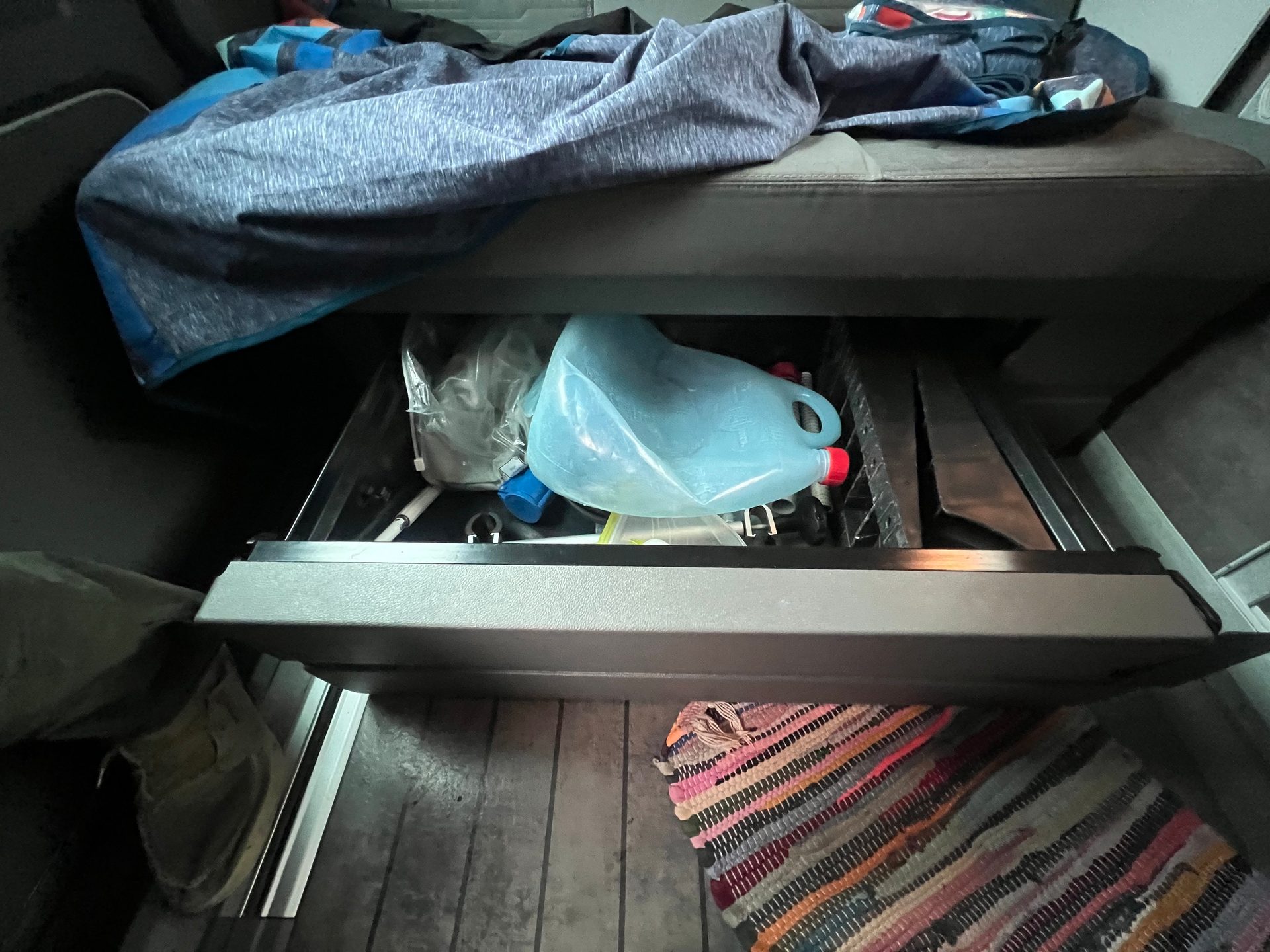 An open storage drawer under a bed contains a blue jug and other items, with a blanket above and a colorful rug on the floor.