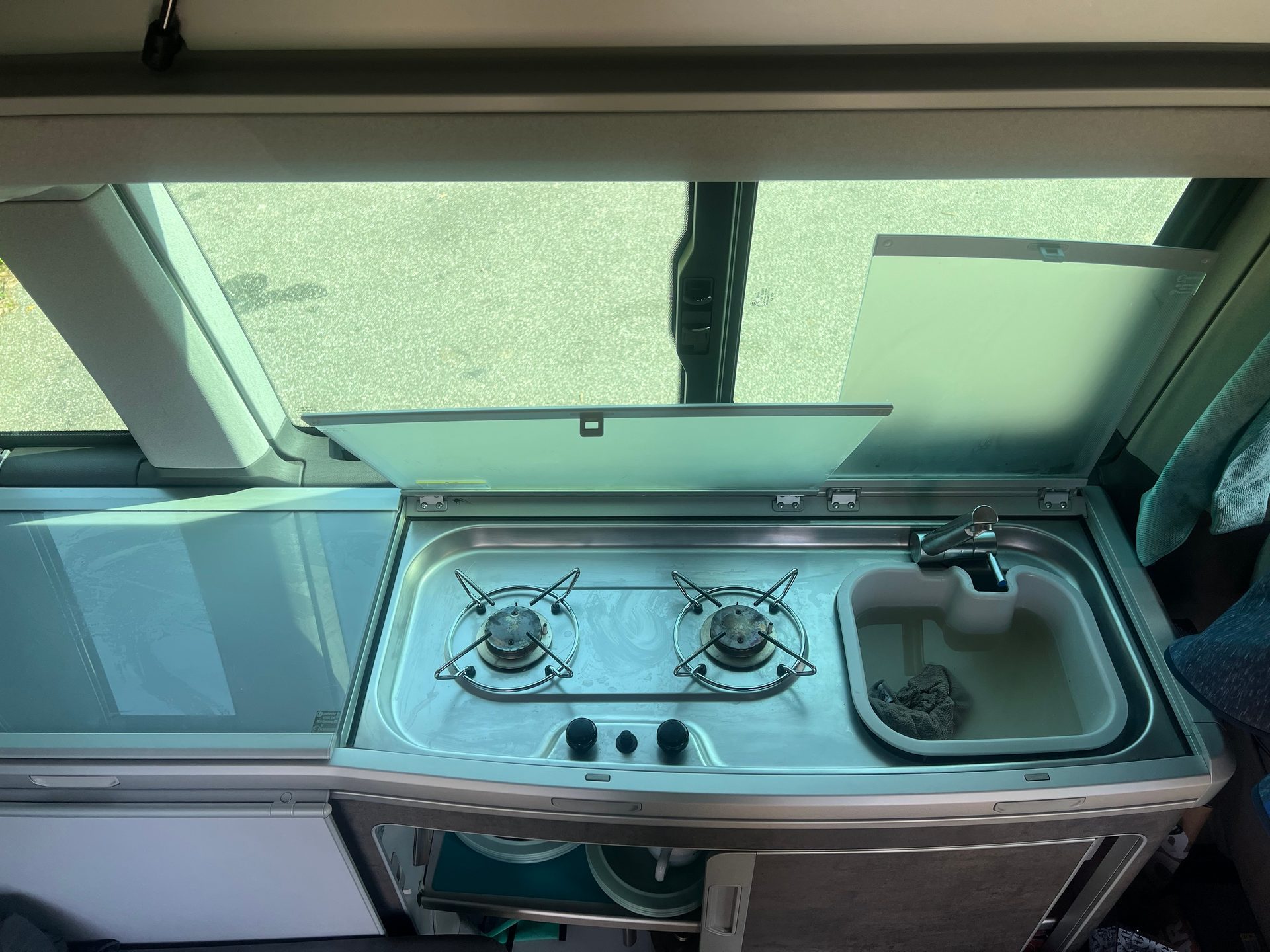 Interior of a camper van kitchen unit with a two-burner stove, sink, and glass cover.