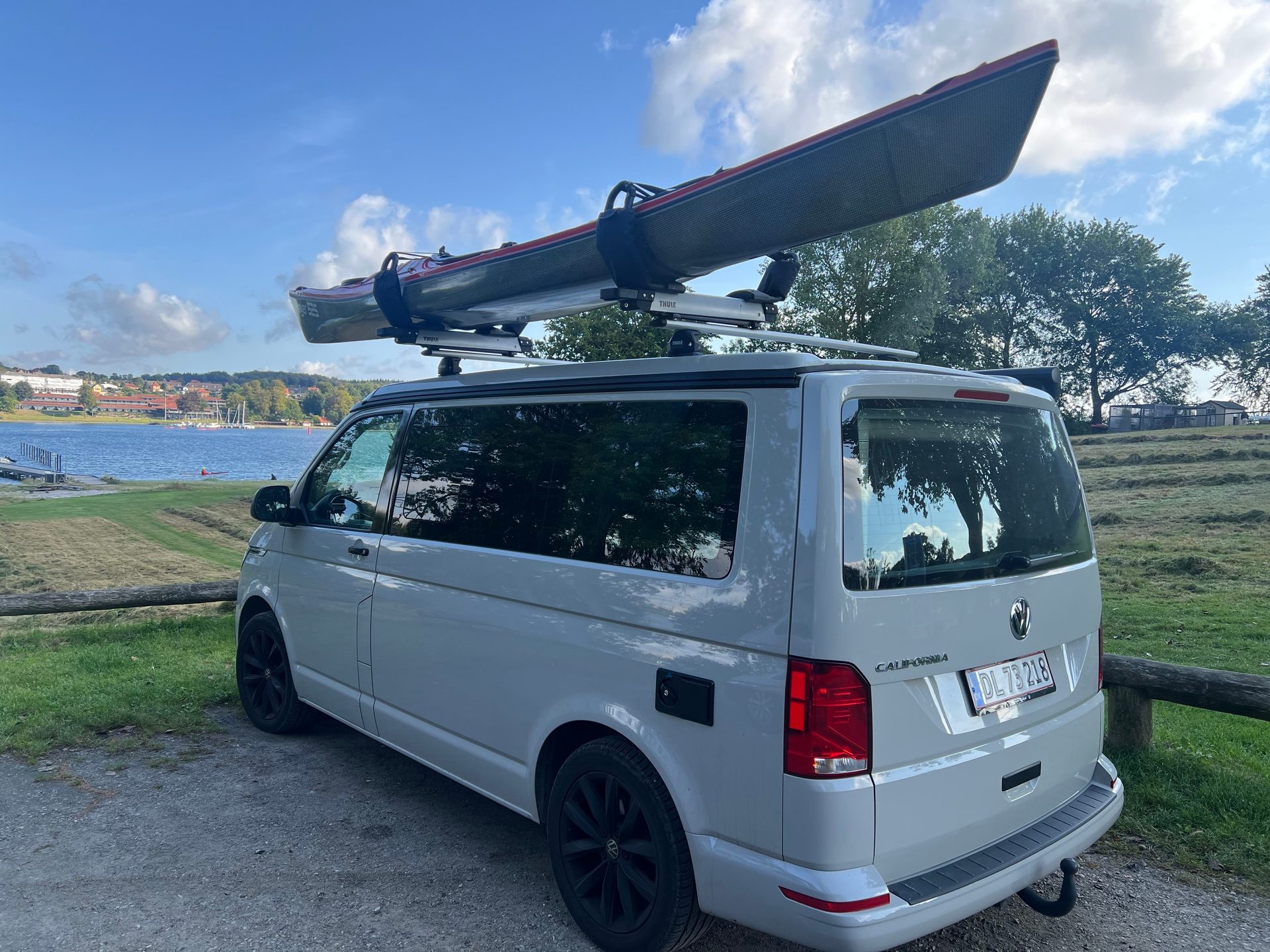 White camper van with a kayak on its roof rack, parked by a lake.